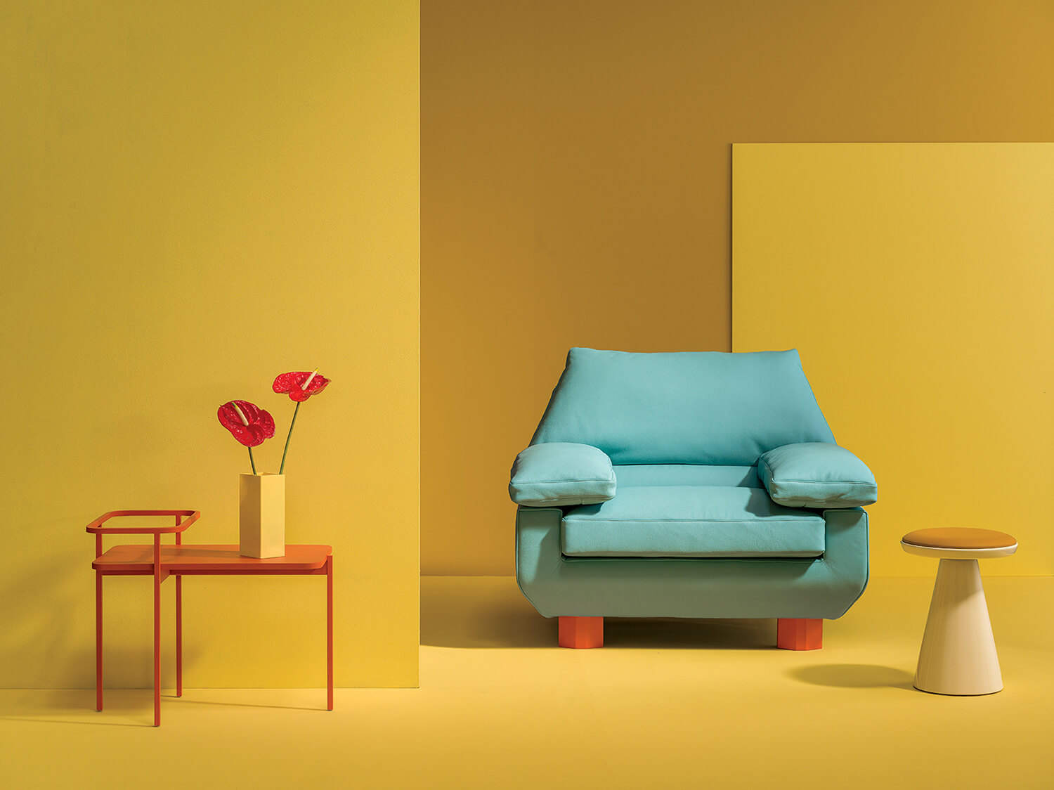 A blue armchair with wooden legs sits between a small orange table holding red flowers and a round white and yellow stool, set against a yellow background.