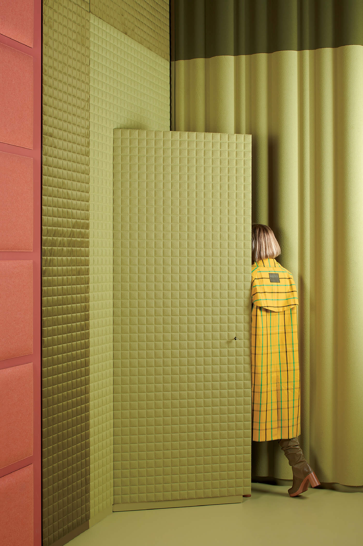 A person in a yellow plaid coat and brown boots stands partially hidden behind a quilted green door in a room with quilted walls and green curtains.
