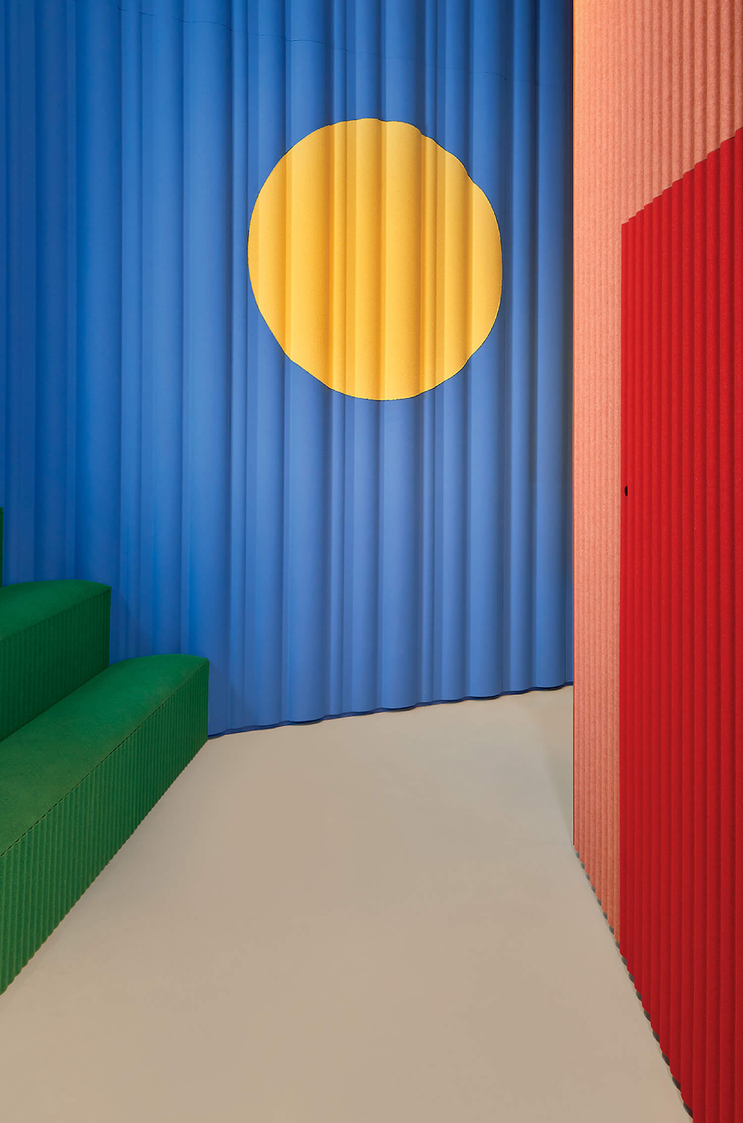 Corrugated blue wall with a large yellow circle, green steps on the left, and a red wall on the right, all with a ribbed texture.