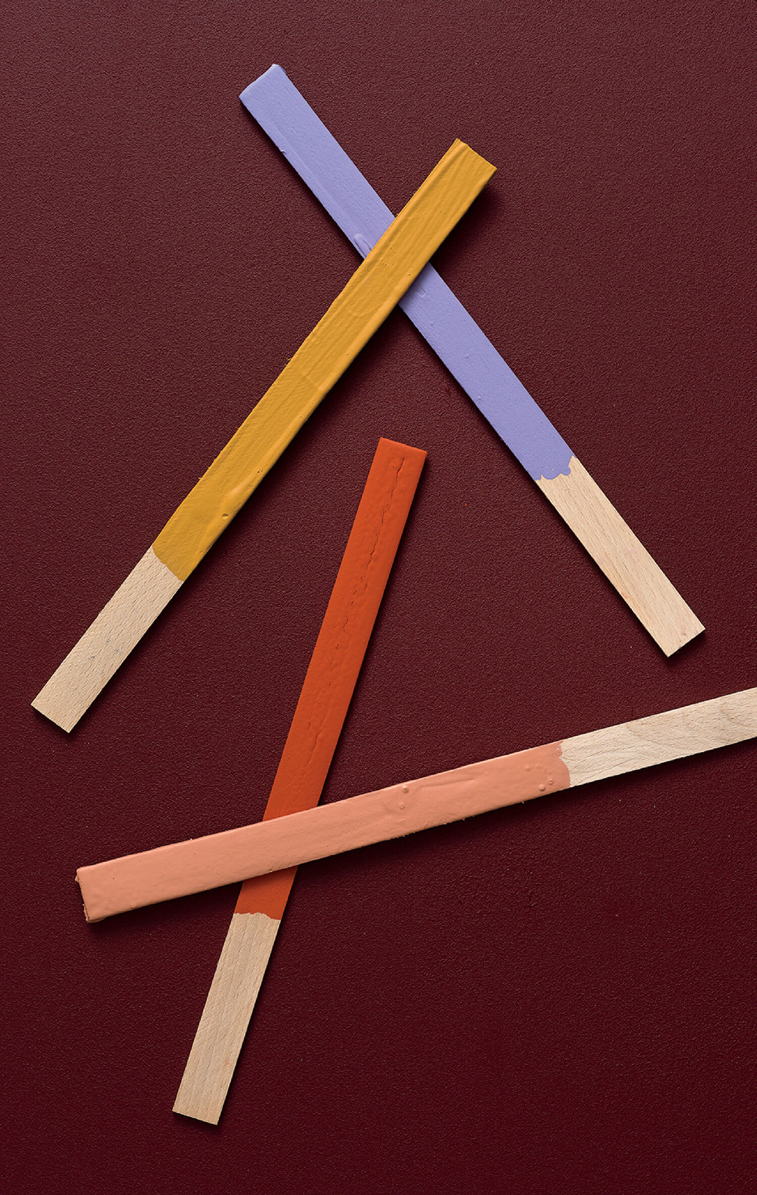 Three wooden paint stirrers, each partly painted in yellow, purple, and orange, are arranged on a dark red textured surface.