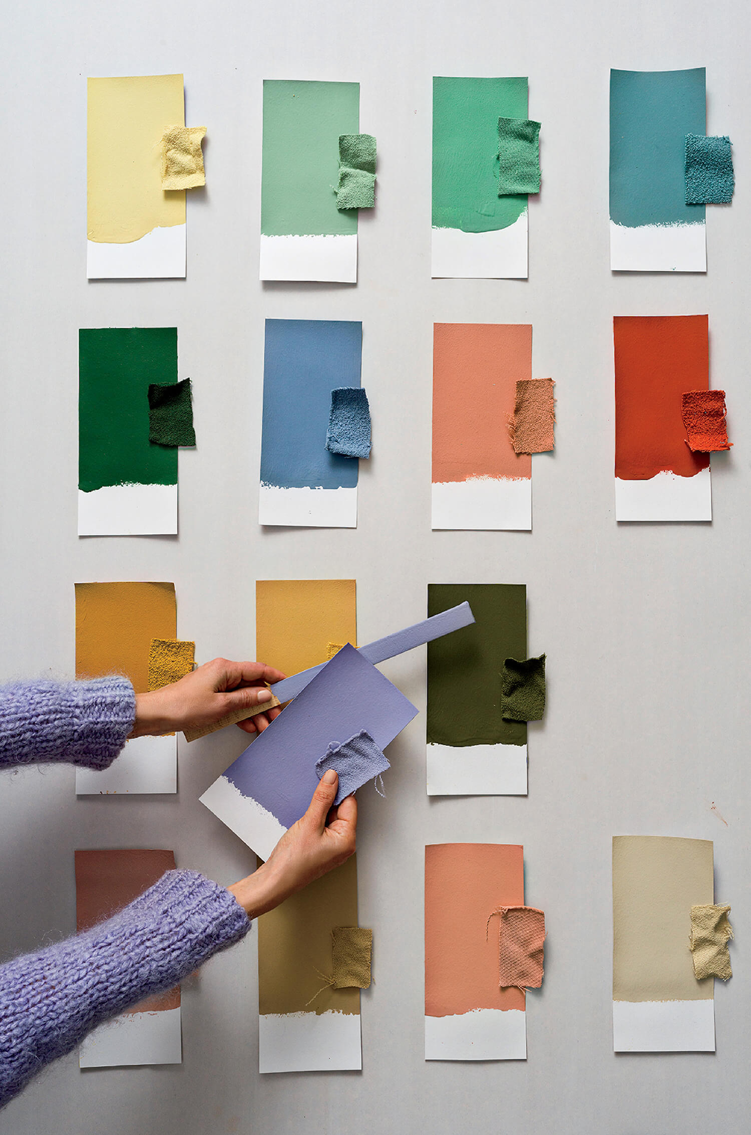 A person holds a lavender paint swatch and fabric sample above a table displaying multiple paint swatches and matching fabric pieces in various colors.