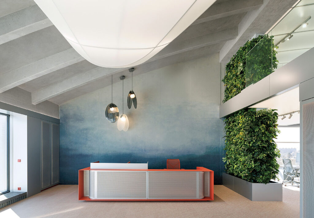 Modern office reception area with a red and white desk, hanging pendant lights, a blue gradient wall, and a vertical green plant wall, with an open workspace visible in the background.