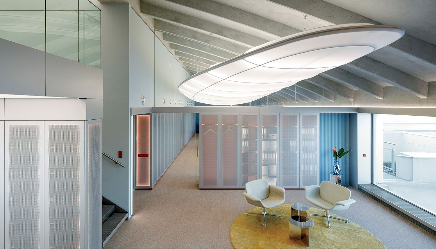 Modern office lounge with two white chairs, a small table on a round yellow rug, glass partitions, and a large curved ceiling light. A staircase is visible to the left.