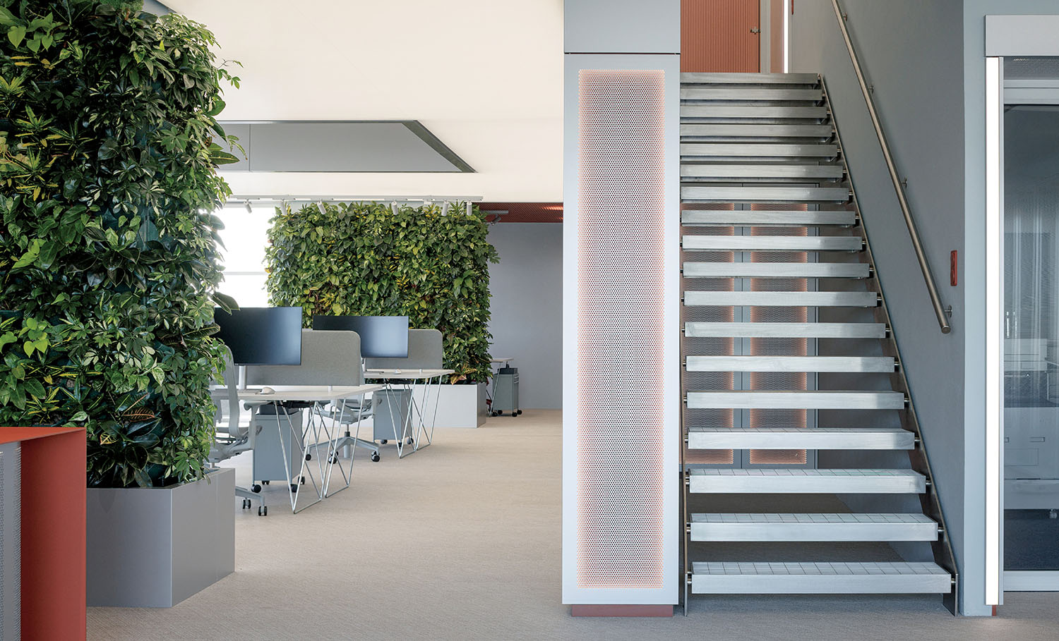 Modern office space with open desks, vertical green walls, and a metal staircase leading to an upper floor.