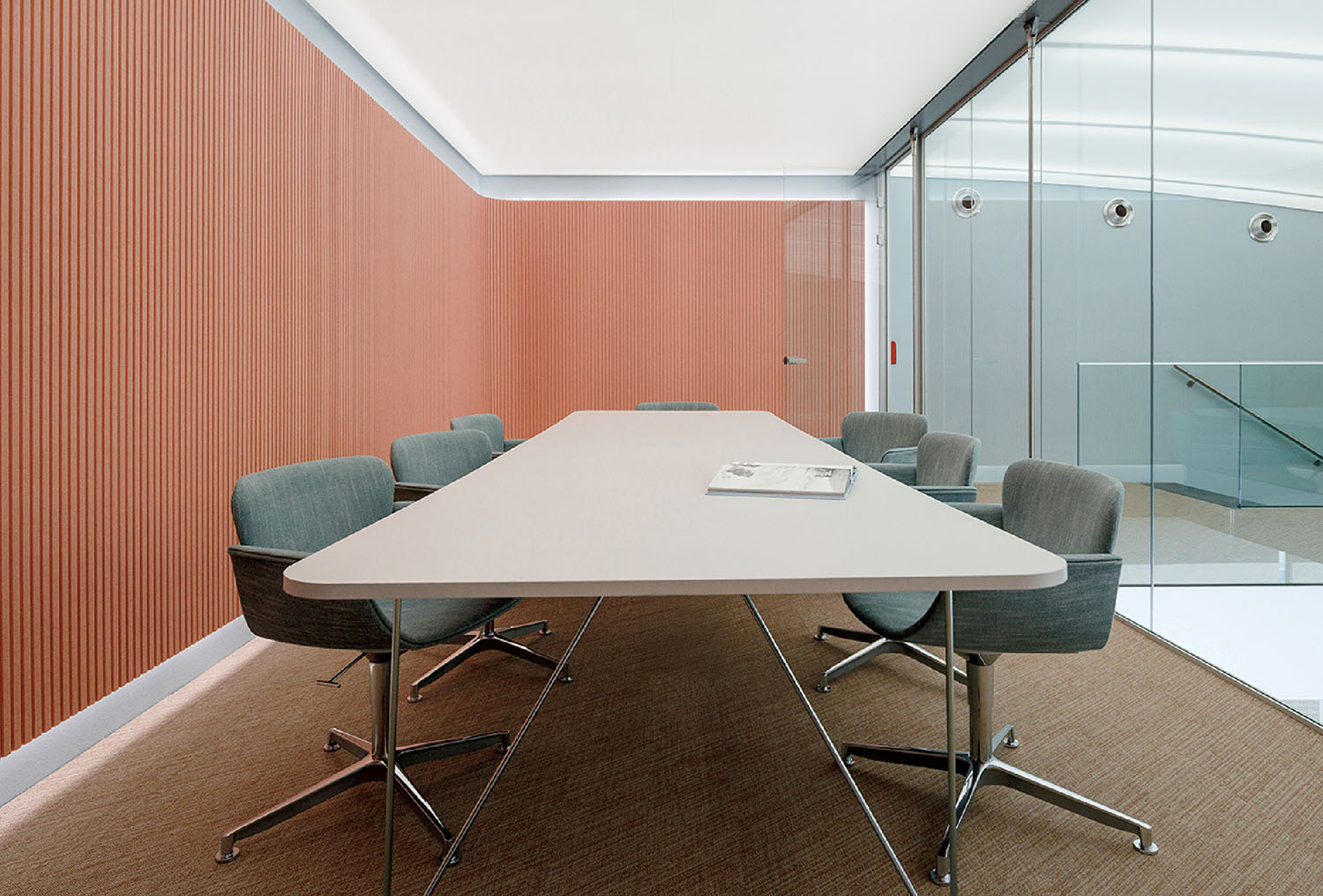 A modern conference room with a long white table, six gray chairs, a magazine on the table, red textured walls, and glass partitions.