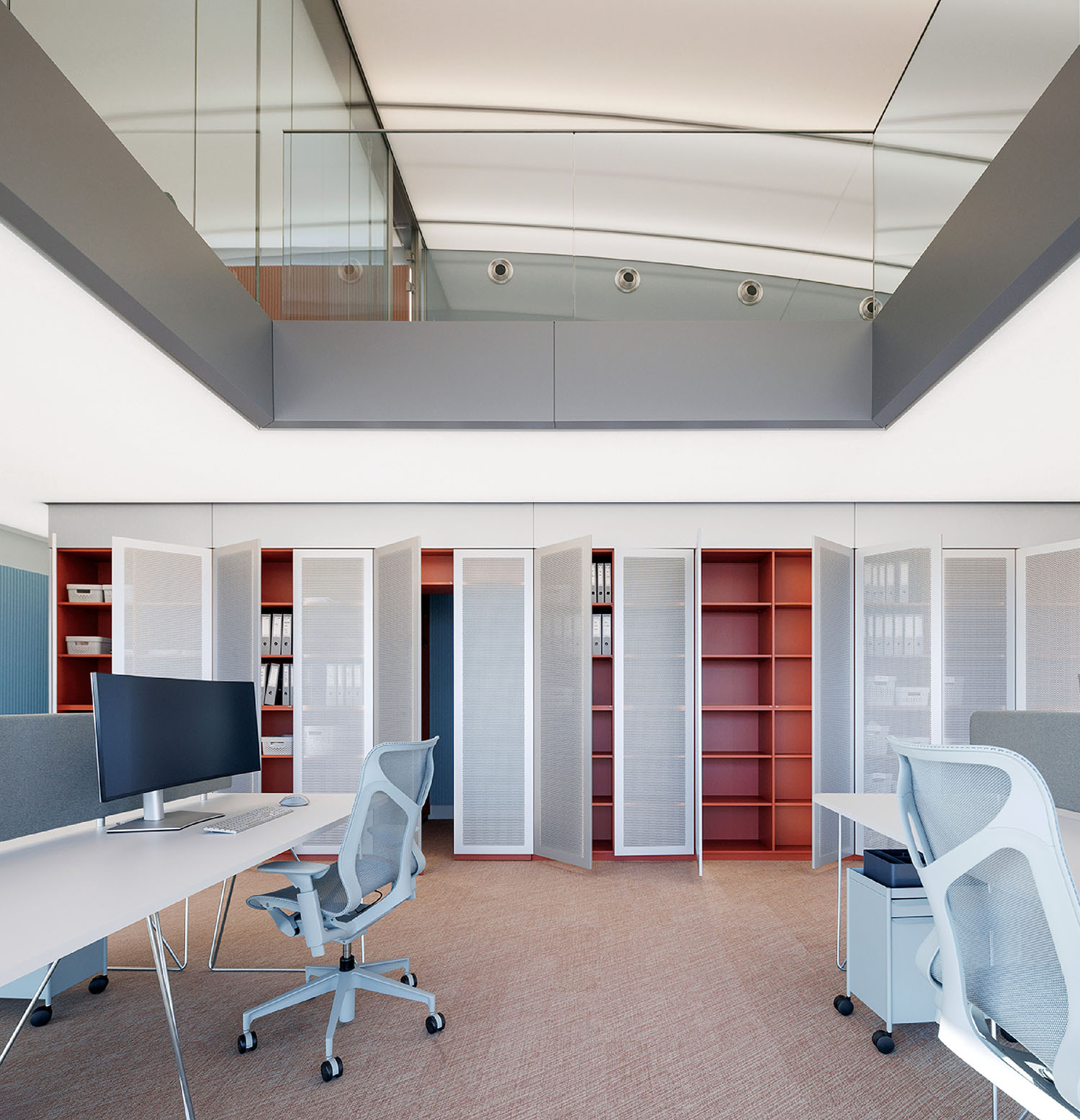 Modern office space with desks, computer monitors, ergonomic chairs, and open shelving units against a wall; upper level with glass railing visible.