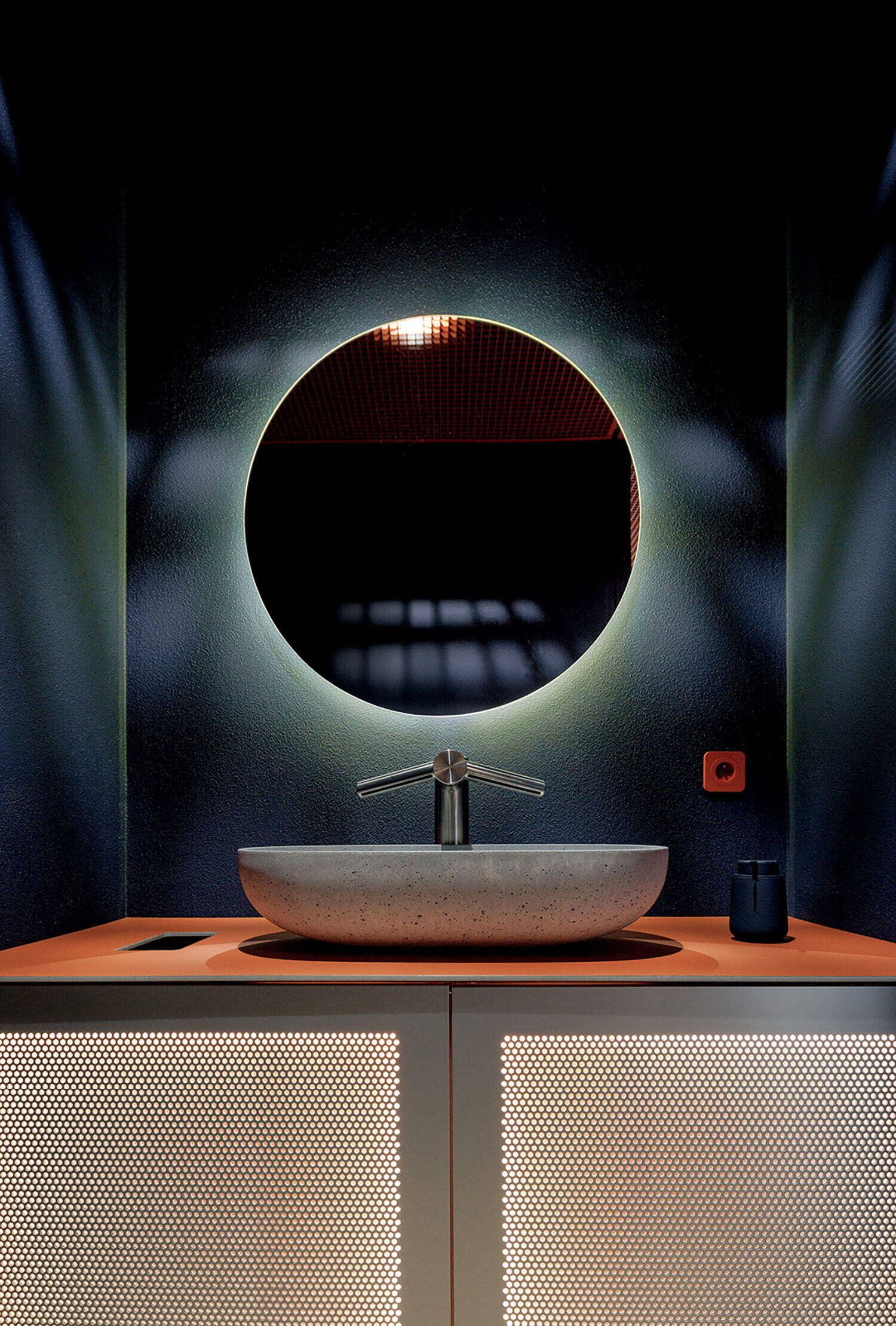Modern bathroom vanity with a round backlit mirror above a oval stone sink, set on a perforated cabinet with a dark background.
