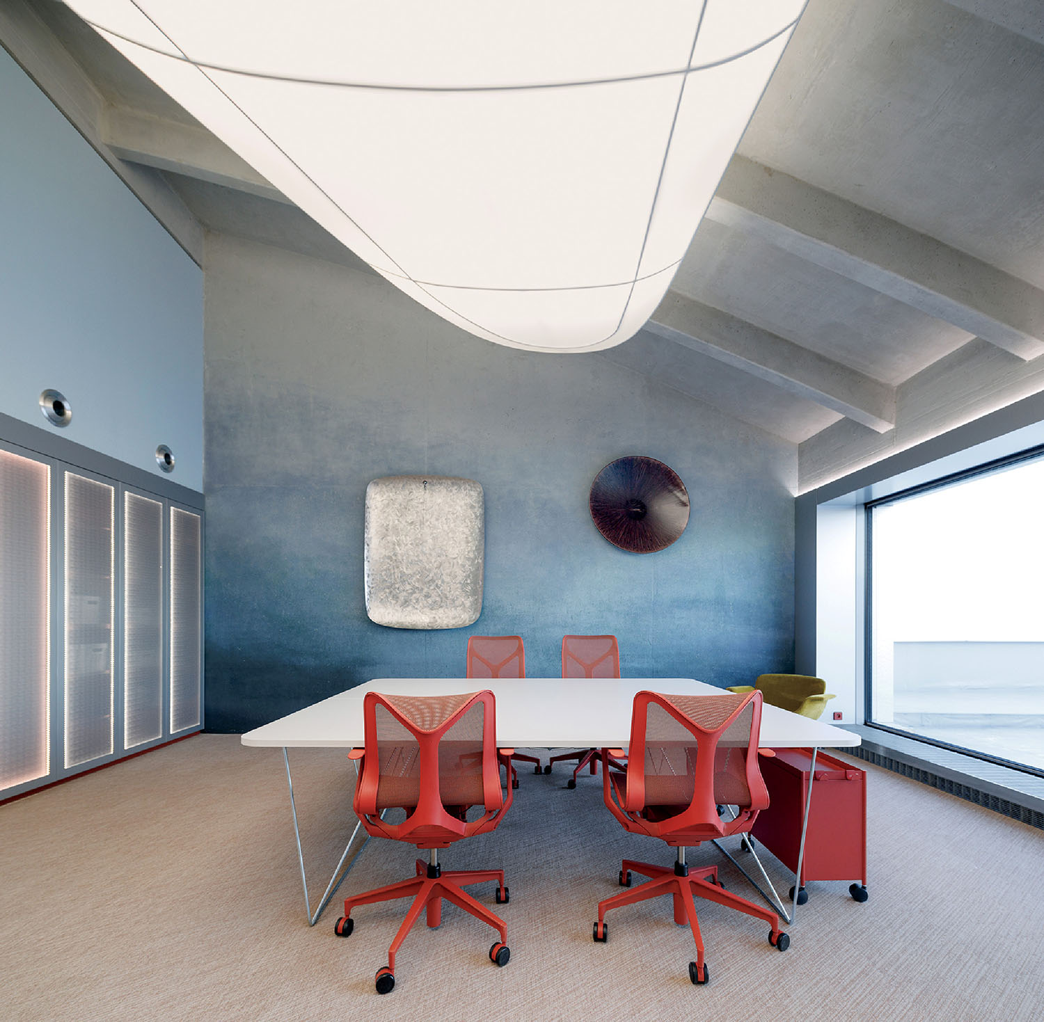 Modern conference room with a rectangular table, four red office chairs, abstract wall art, large overhead light, and floor-to-ceiling window providing natural light.