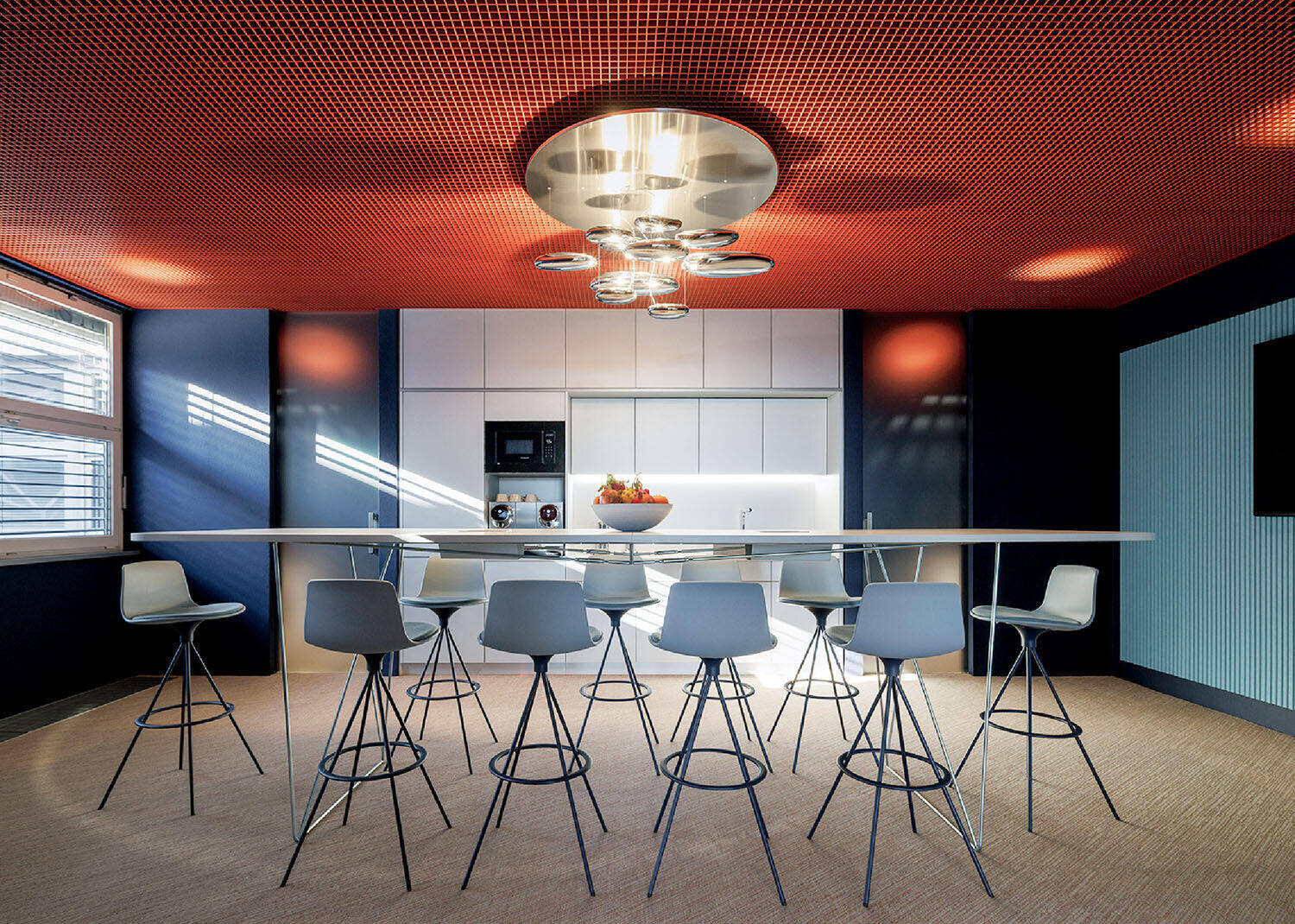 Modern kitchen with a long white table, nine gray bar stools, minimalist white cabinets, and a metallic ceiling light fixture. Natural light enters through two large windows.