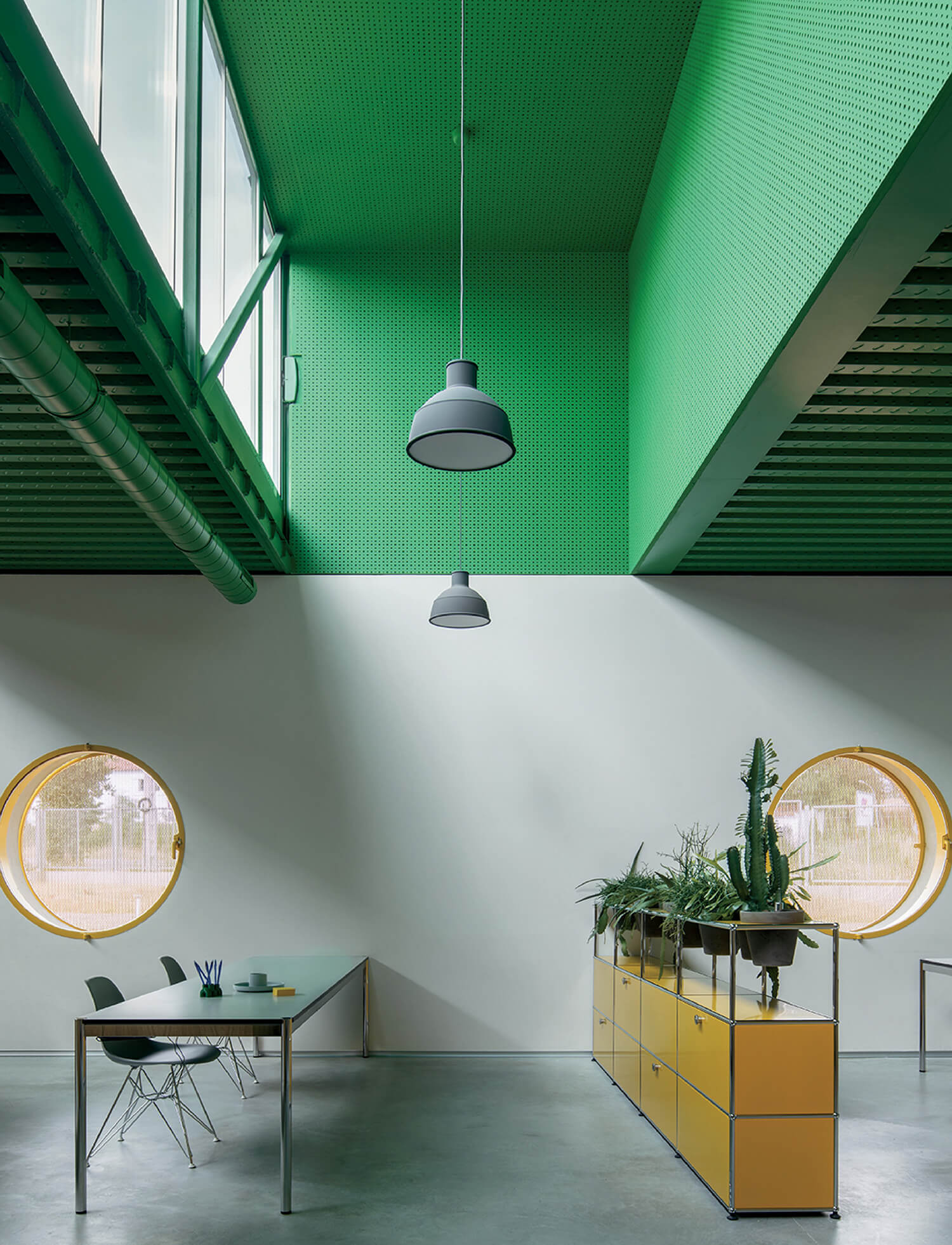 Modern office space with green ceiling, large windows, round yellow-framed windows, a table, and yellow storage unit topped with potted plants.
