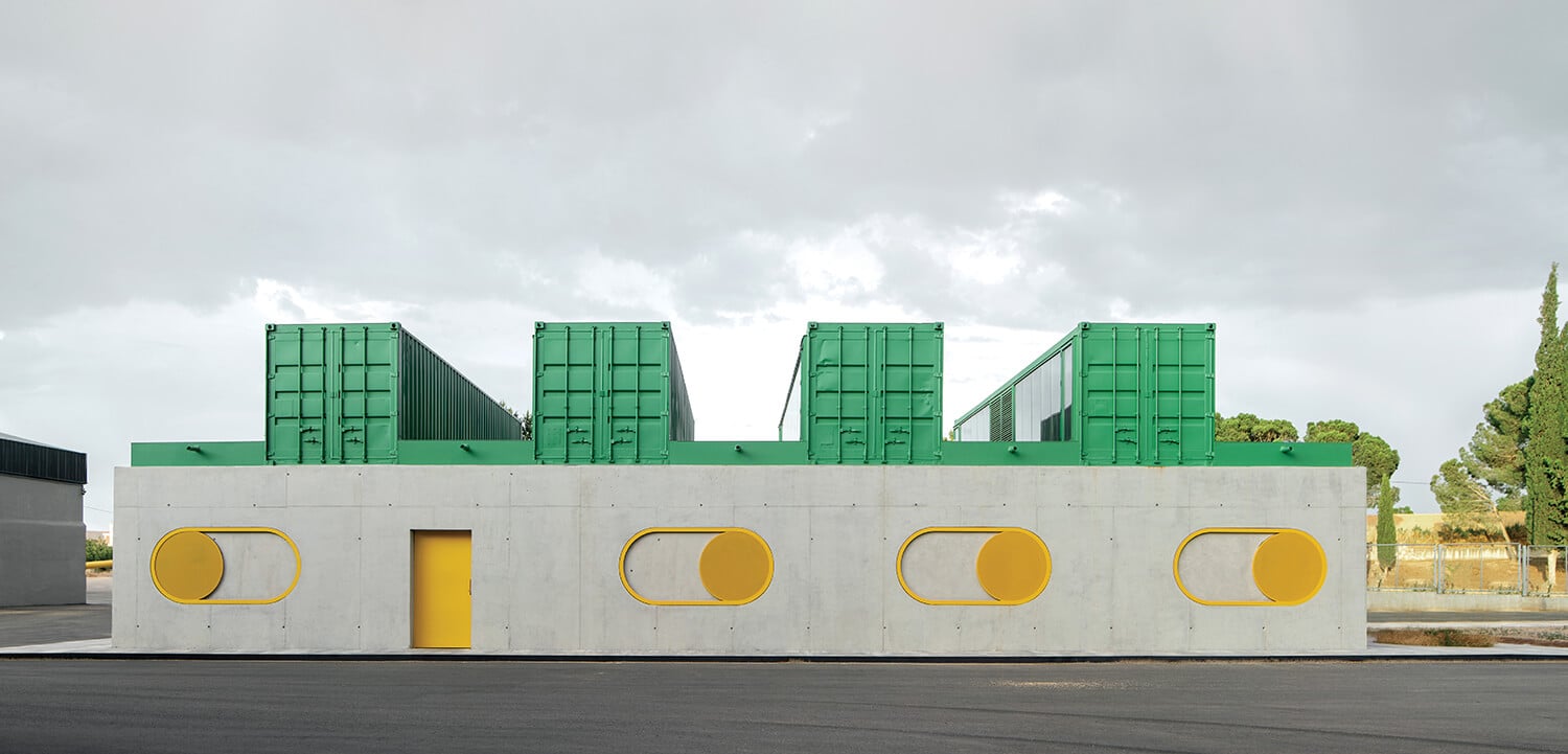 A modern, rectangular building with a gray concrete facade, yellow door and window frames, and green shipping containers stacked on the roof.