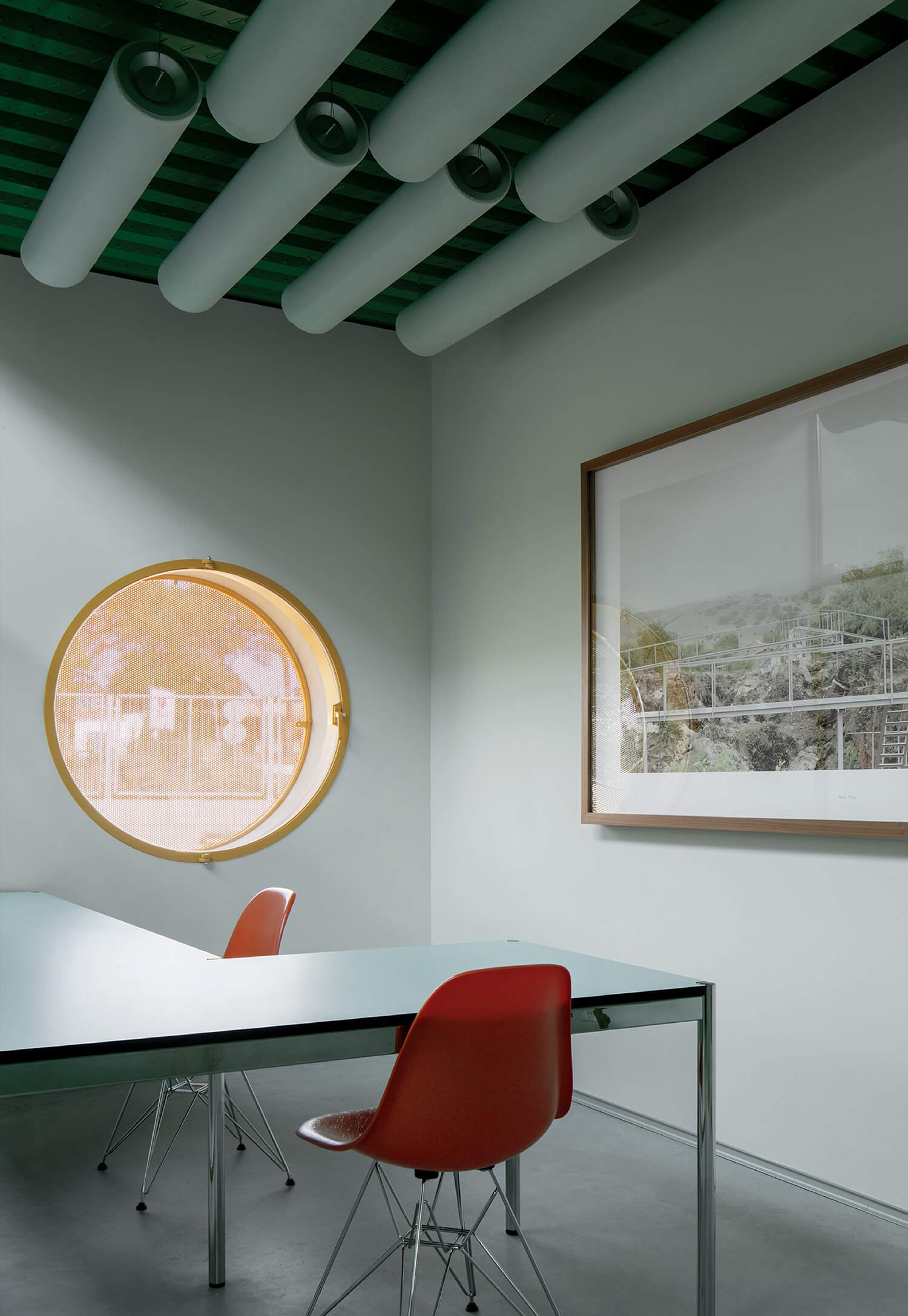 Minimalist office with a round window, two red chairs, a glass table, and a large framed photograph on the wall. Ceiling features exposed cylindrical ducts.