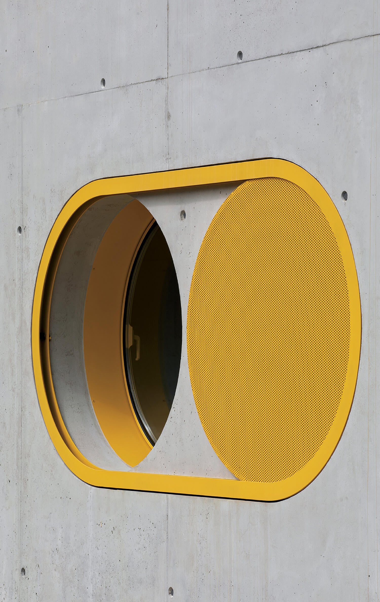 Yellow-rimmed oval window set in a concrete wall, partially covered by a yellow perforated metal panel.