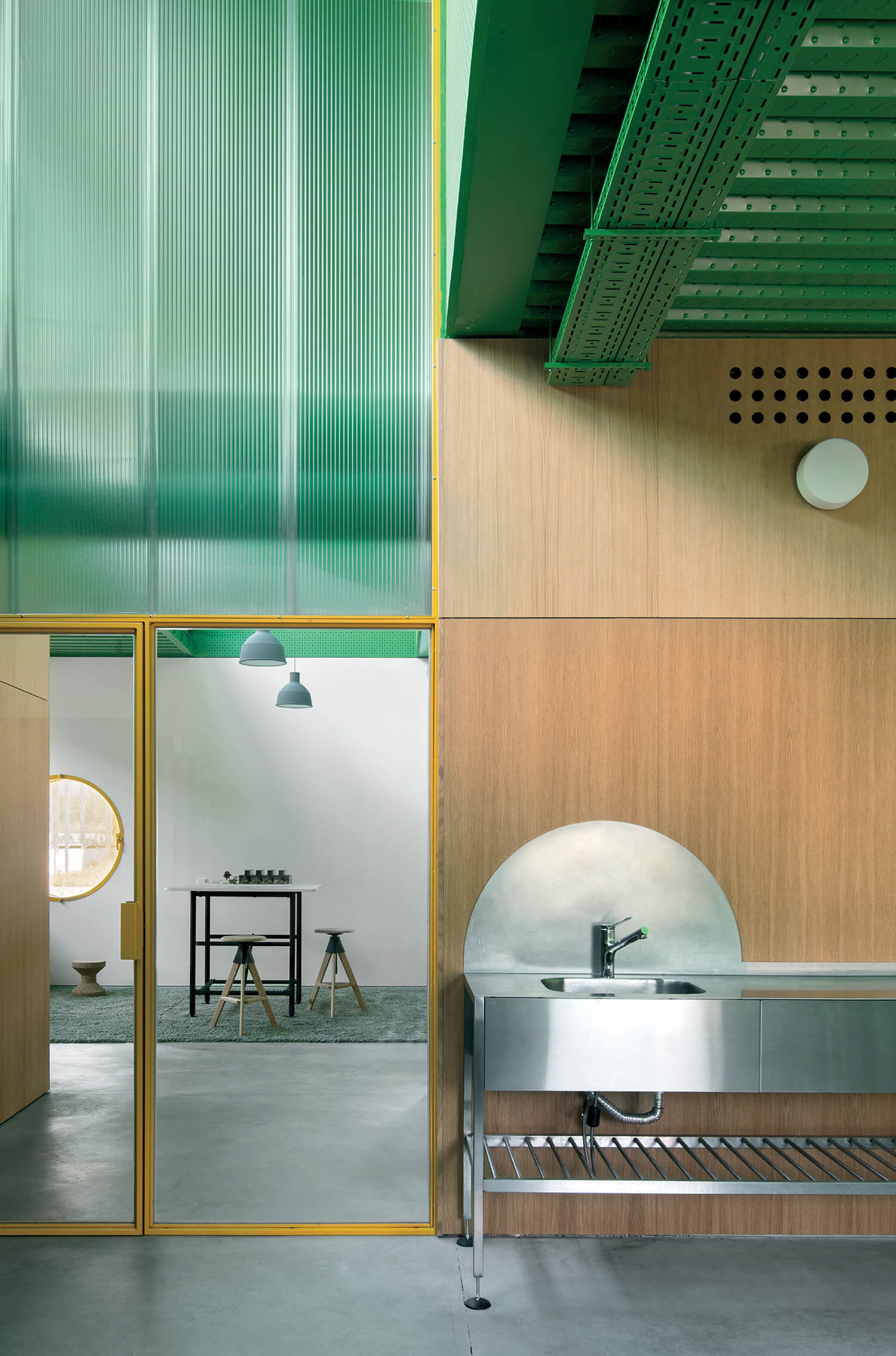 Modern kitchen interior with a stainless steel sink, wood panel walls, yellow-trimmed glass door, and exposed green ceiling beams. A minimal white room with stools is visible through the door.