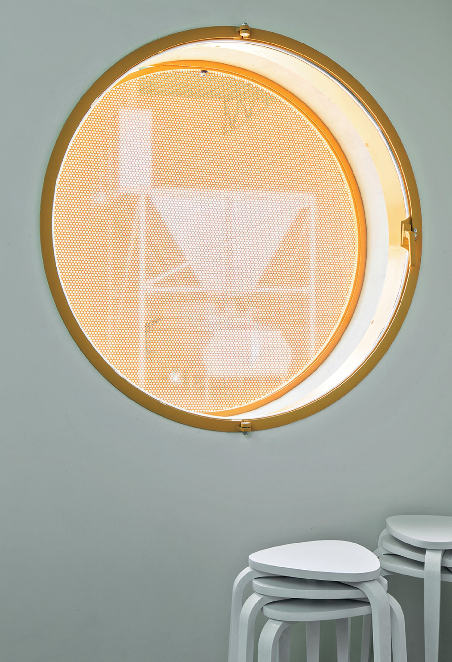 A round, yellow-framed window with a mesh screen, revealing industrial equipment outside; three stacked white stools sit below the window.