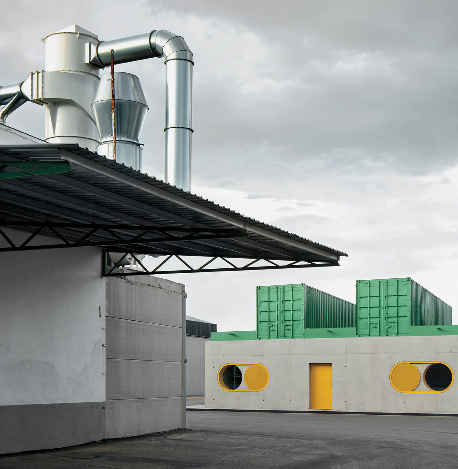 Industrial facility with exposed metal ducts, a corrugated roof, and a modern building featuring green containers and circular yellow windows and door. Overcast sky in the background.