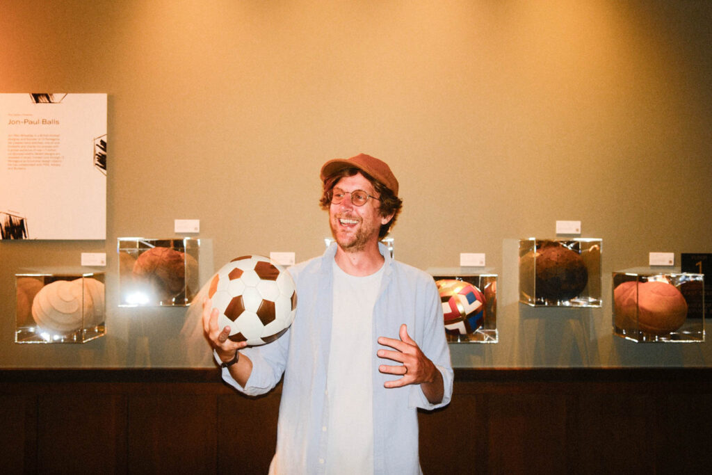 A man wearing glasses and a cap smiles while holding a soccer ball, standing in front of a display of various historical soccer balls in glass cases.