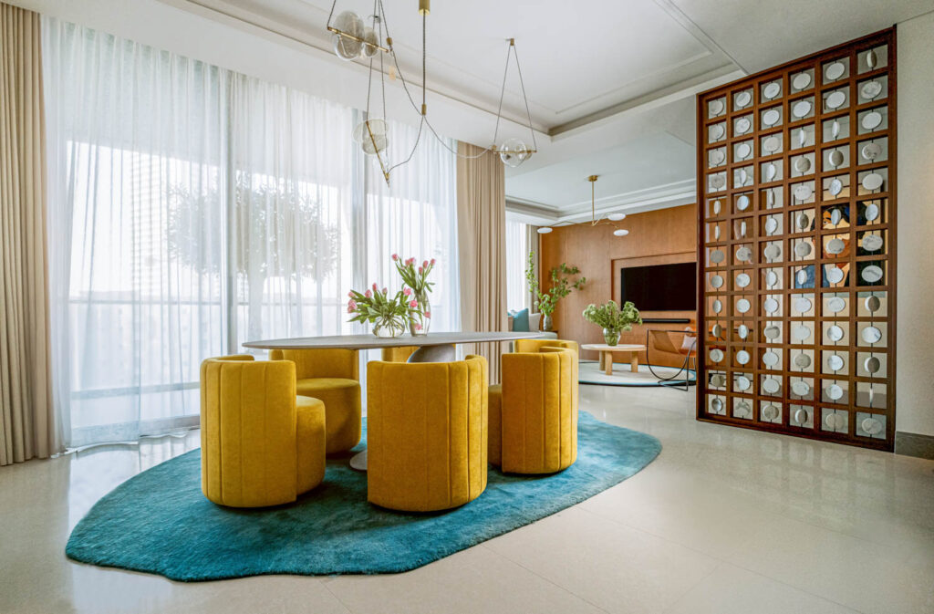 Modern dining area with a round table, six yellow chairs, blue rug, large windows with sheer curtains, and a partial room divider next to a living room with a TV.
