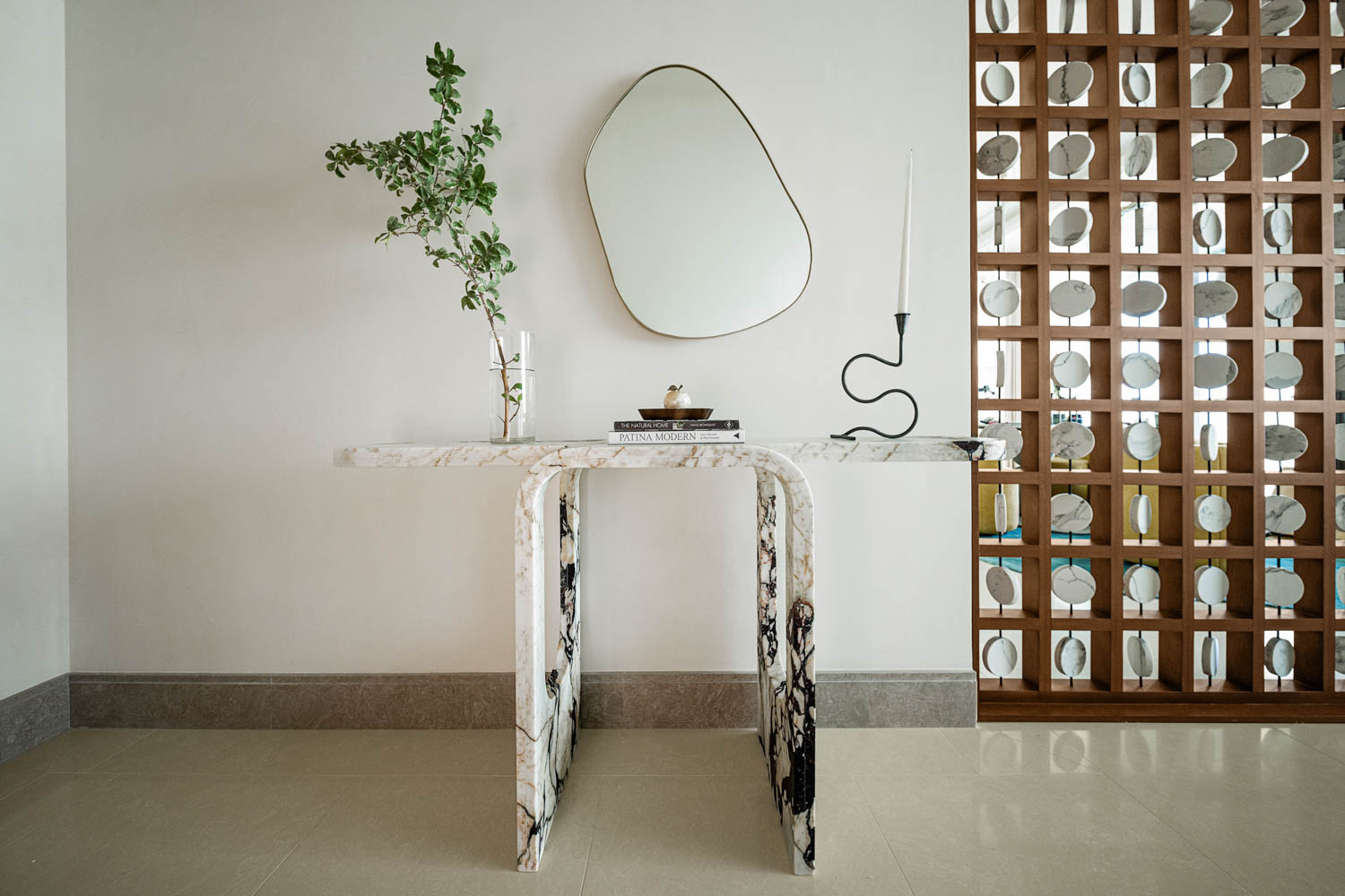 A marble console table with a vase, books, a decorative object, and a candle stands against a white wall with an abstract mirror above and a wooden partition beside it.