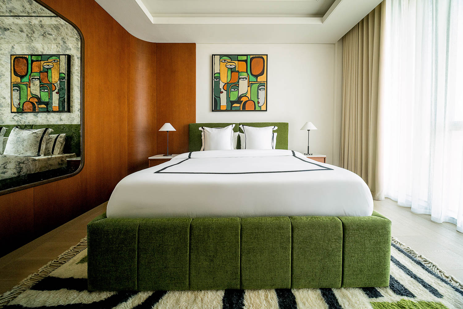 Modern bedroom with a green upholstered bed, white bedding, abstract art above the headboard, and large window with sheer curtains.