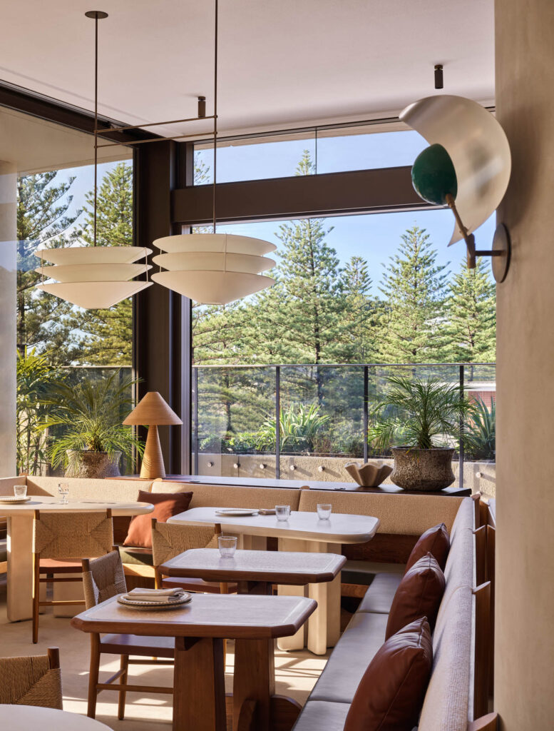 Modern restaurant interior with wooden tables, cushioned seating, large windows, pendant lights, and views of trees outside.