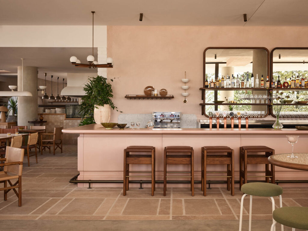 A modern restaurant bar with light wood decor, a pink counter, wooden stools, shelves with bottles, and large mirrors behind the bar. Tables and chairs are arranged throughout the space.