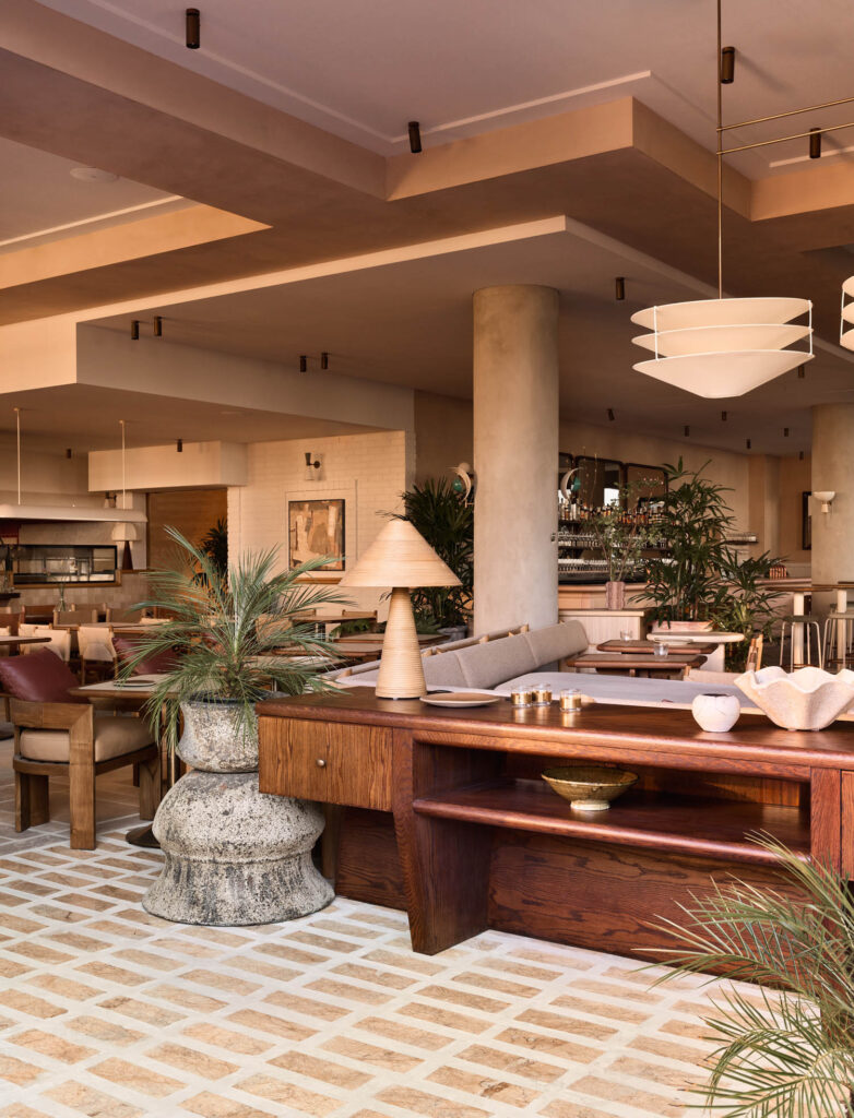 Modern restaurant interior with wooden furniture, potted plants, soft lighting, and neutral tones. Large light fixtures hang from the ceiling above the seating area.