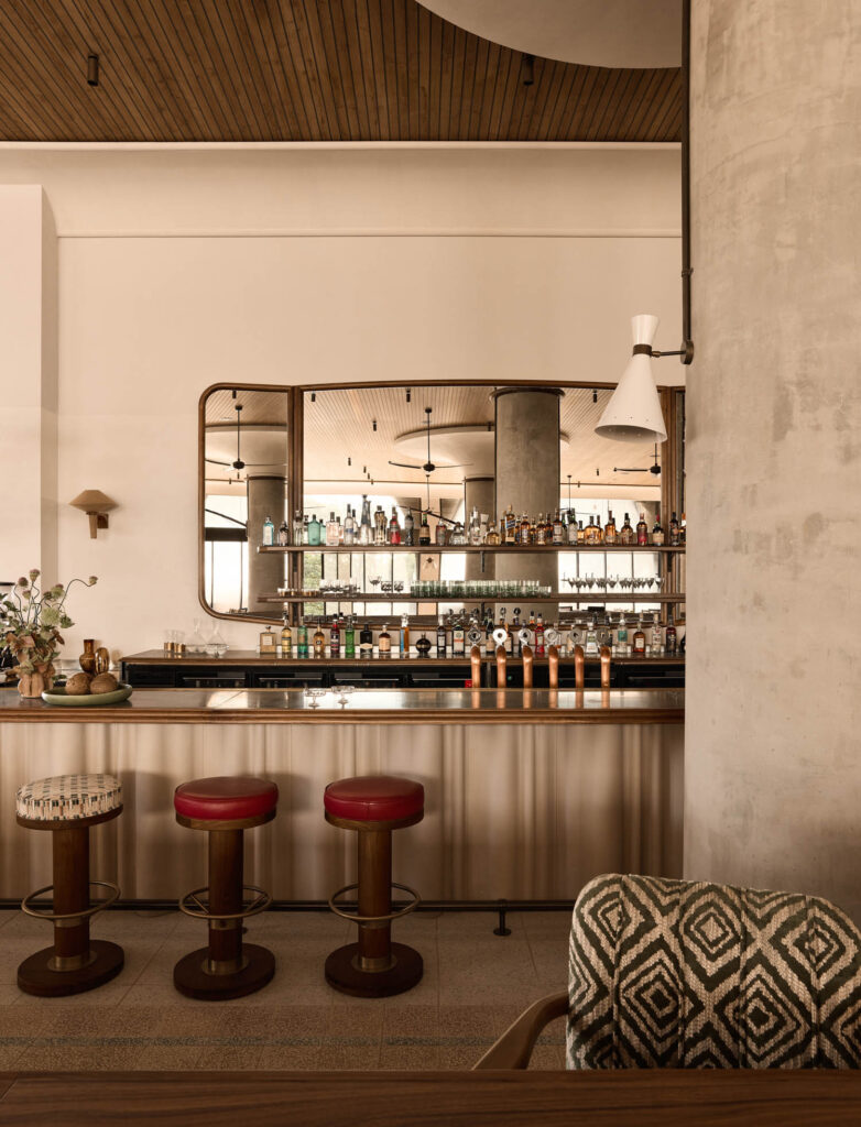 Modern bar interior with red-cushioned stools, a patterned chair, large wall mirror, shelves of bottles, and neutral-toned decor.