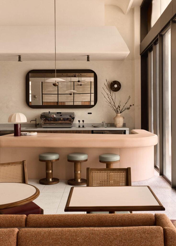 Minimalist café interior with light pink curved counter, two stools, a coffee machine, large wall mirror, vase with branches, and tables with chairs near floor-to-ceiling windows.