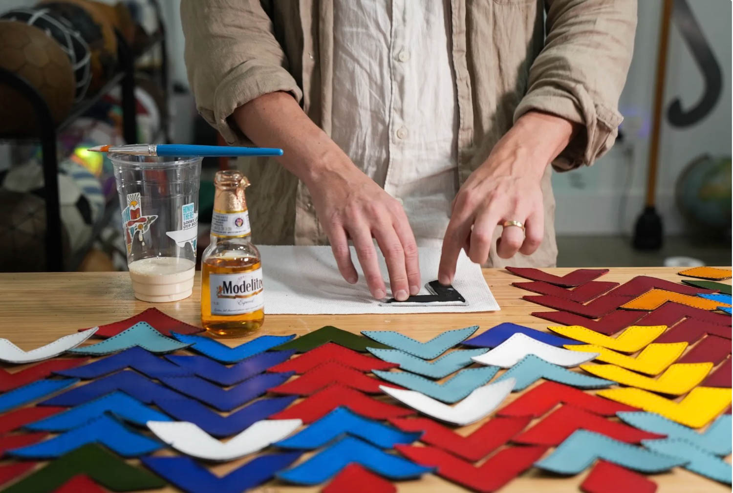 A person arranges colored leather pieces on a table with a paintbrush, plastic cup, and a bottle of Modelo beer nearby.