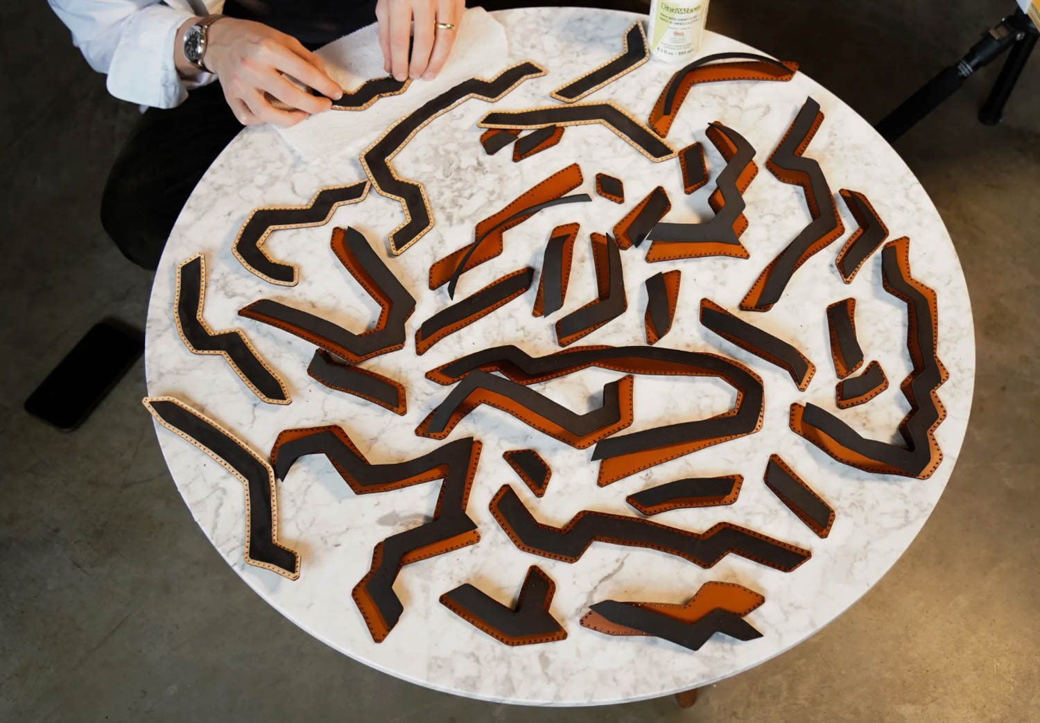 A person arranges several irregularly shaped, stitched brown leather pieces on a round white marble table.