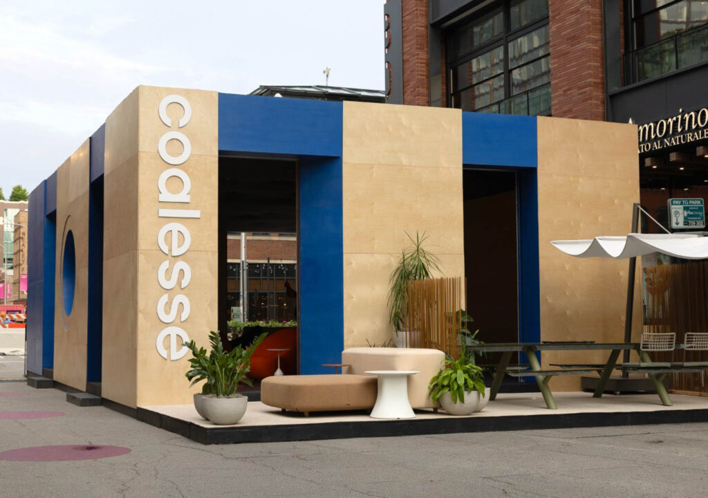 Modern outdoor pavilion with "Coalesse" signage, beige and blue exterior panels, contemporary seating, plants, and picnic tables, situated near shops in an urban setting.