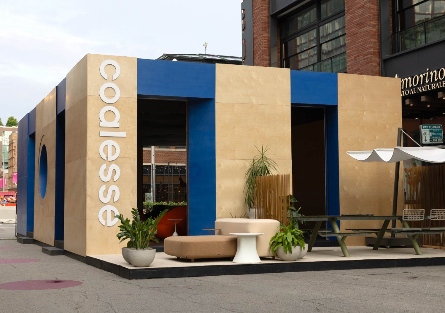 Modern outdoor pavilion with "Coalesse" signage, beige and blue exterior panels, contemporary seating, plants, and picnic tables, situated near shops in an urban setting.