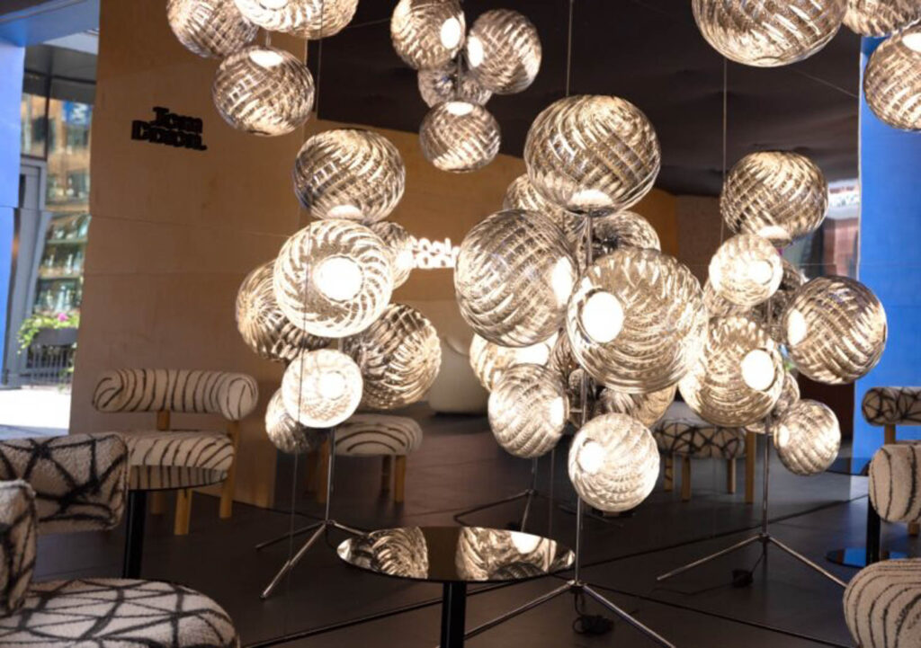 Cluster of spherical pendant lights with striped patterns hangs above a round mirror table, surrounded by upholstered chairs in a modern interior space.