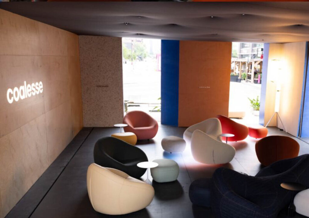 Modern lounge area with curved chairs and small tables, illuminated by natural light from large windows; "coalesse" is projected on the wall.
