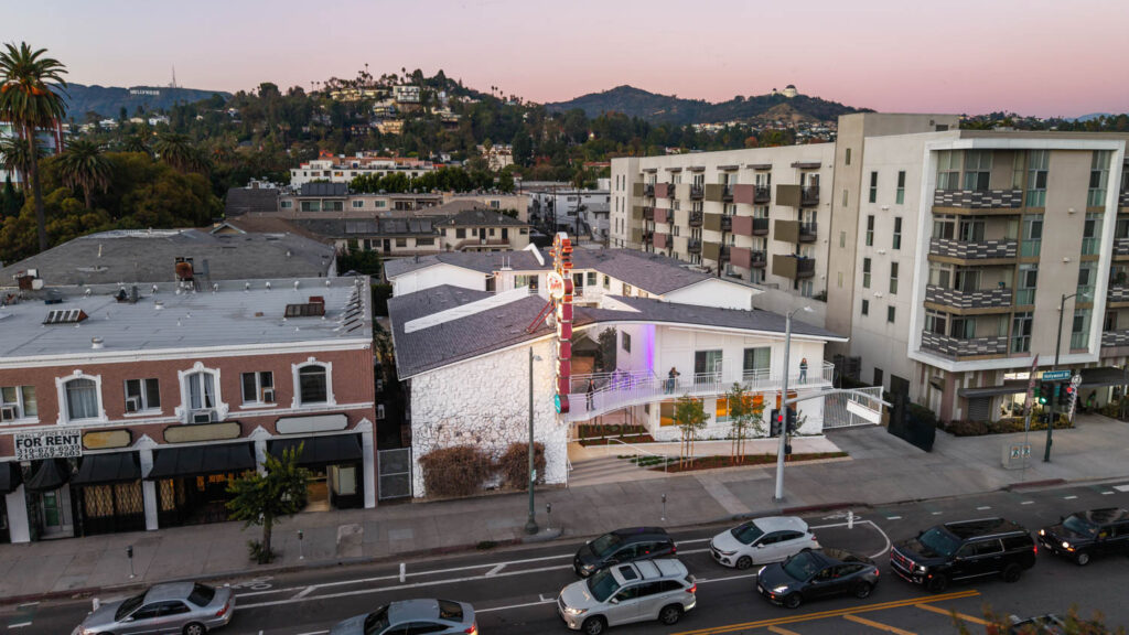 Aerial view of a mid-century motel on a busy street, surrounded by apartments and hills in the background at dusk.