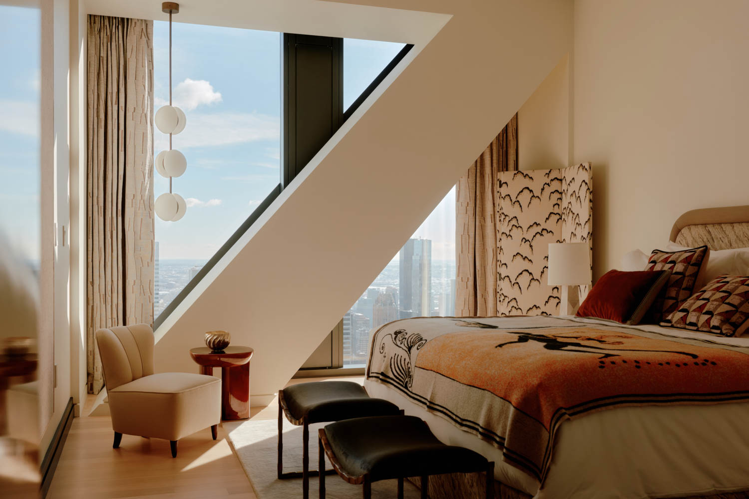 Modern bedroom with geometric window, city view, bed with orange blanket, two black benches, armchair, side table, and floor lamp with globe lights.