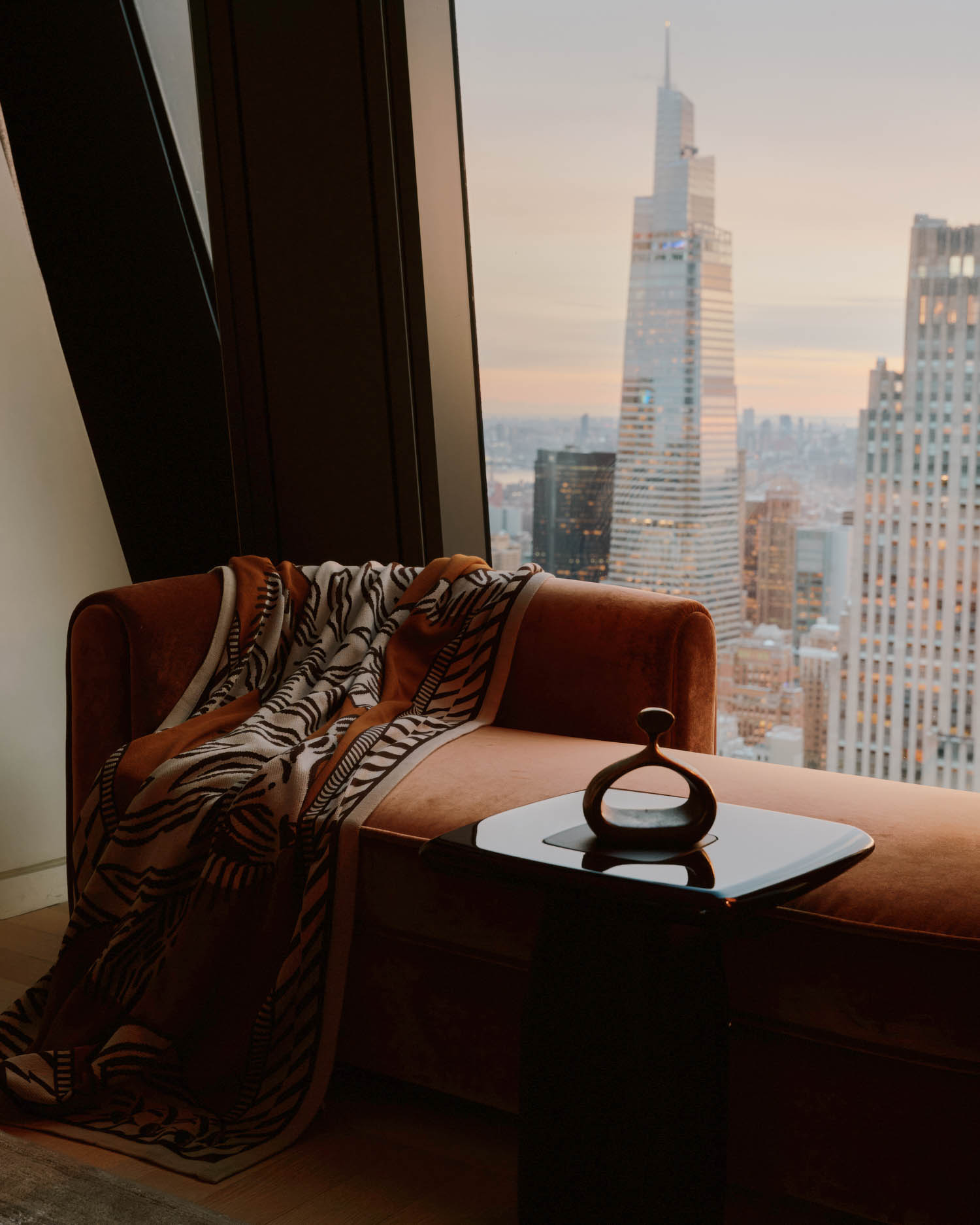 A patterned blanket drapes over a brown velvet sofa beside a small black table, with a city skyline and tall building visible through the window at sunset.
