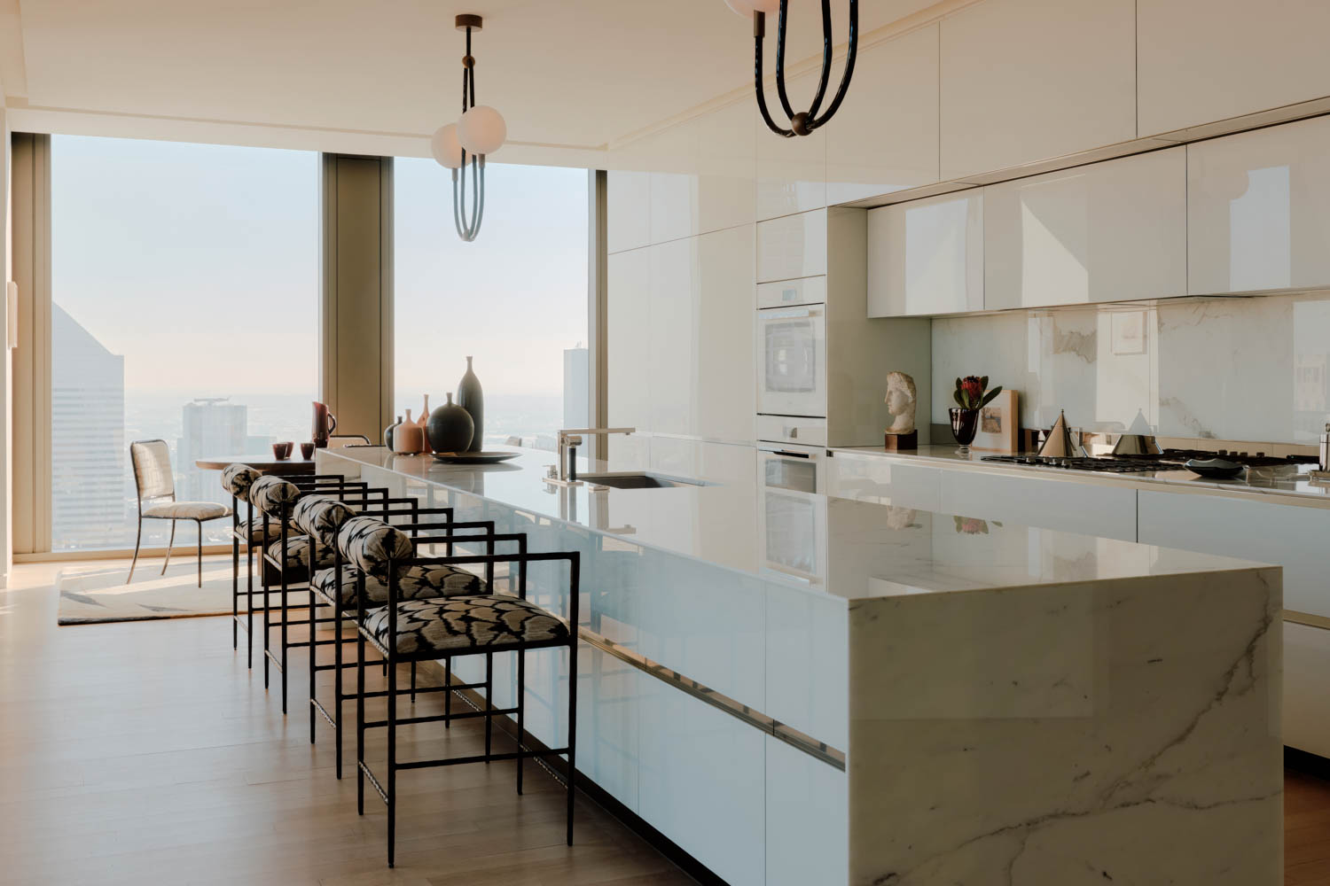 Modern kitchen with white marble island, minimalist cabinetry, four patterned barstools, built-in appliances, and large windows showing a city view.
