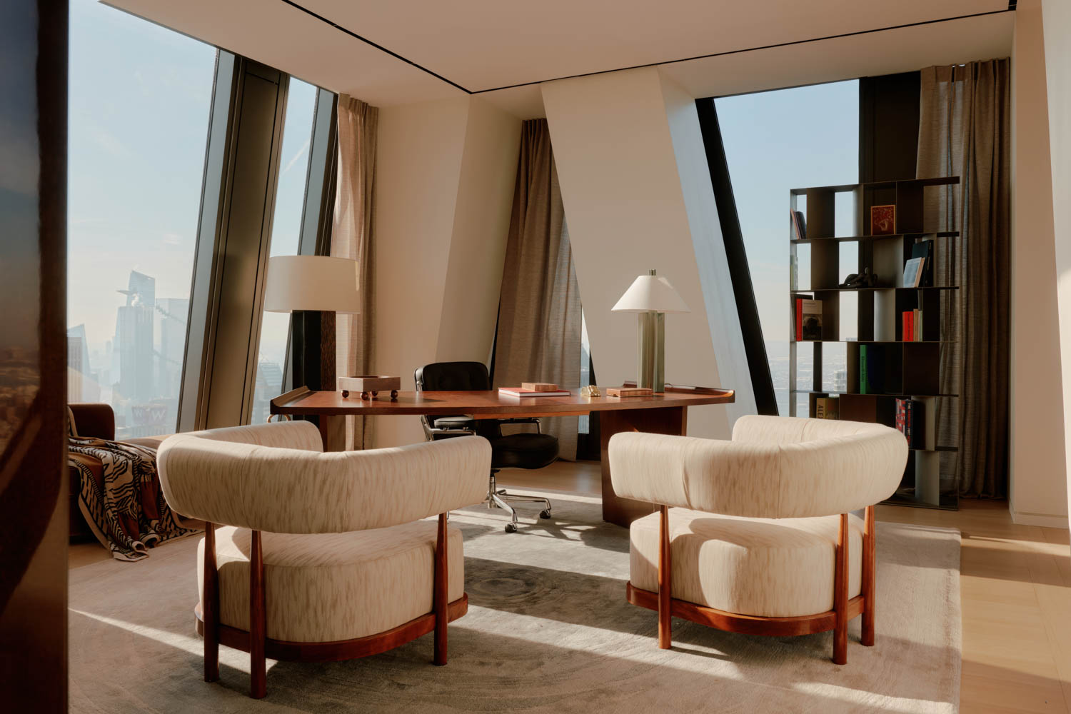Modern office with large windows, a wooden desk, two cream armchairs, shelves, and city buildings visible outside. Natural light fills the room, creating a bright and clean atmosphere.