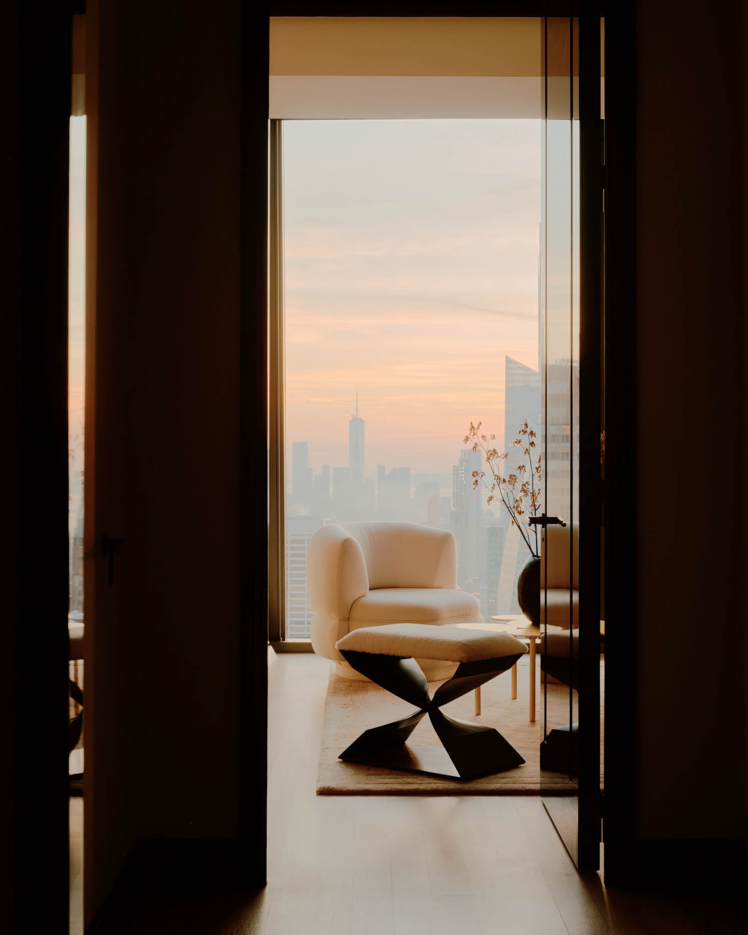 A modern living space with a white armchair, a black coffee table, and a tall window showing a city skyline at sunset.