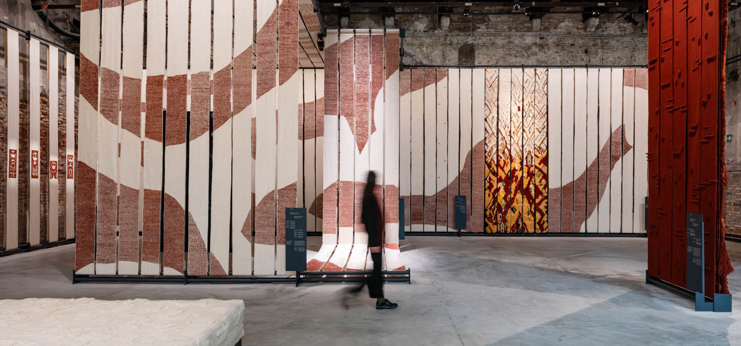 A person walks past large vertical panels featuring abstract red and cream textile patterns in a spacious, industrial-style exhibition hall.