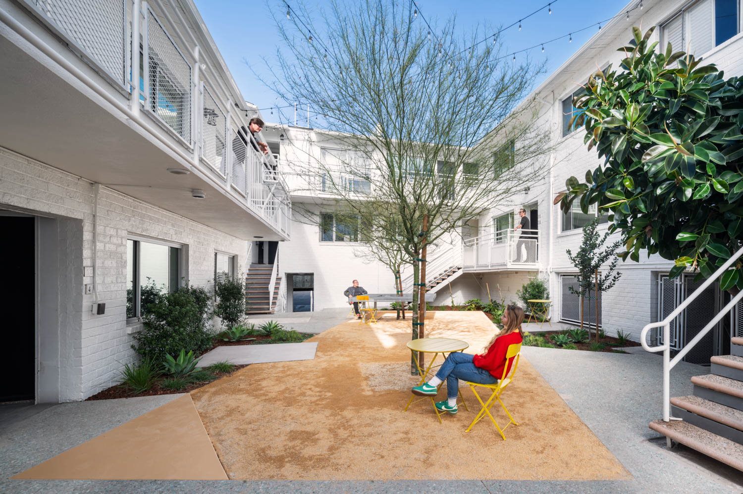 A courtyard with yellow outdoor furniture, a tree in the center, two people seated, one person on a balcony, and white apartment buildings surrounding the area.