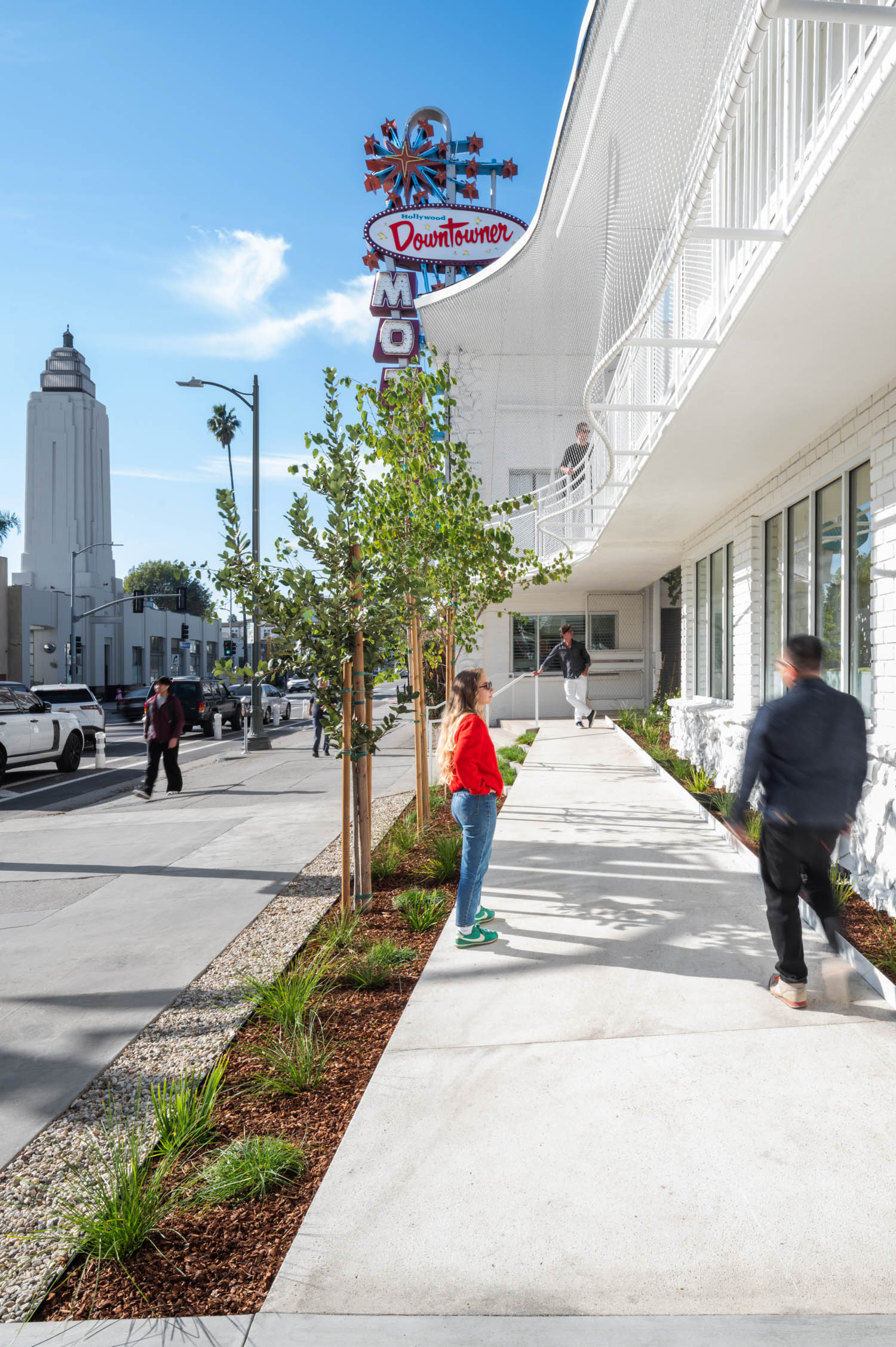 People walk on a wide sidewalk next to a white building with a “Downtowner” motel sign; a tall white tower is visible in the background under a blue sky.