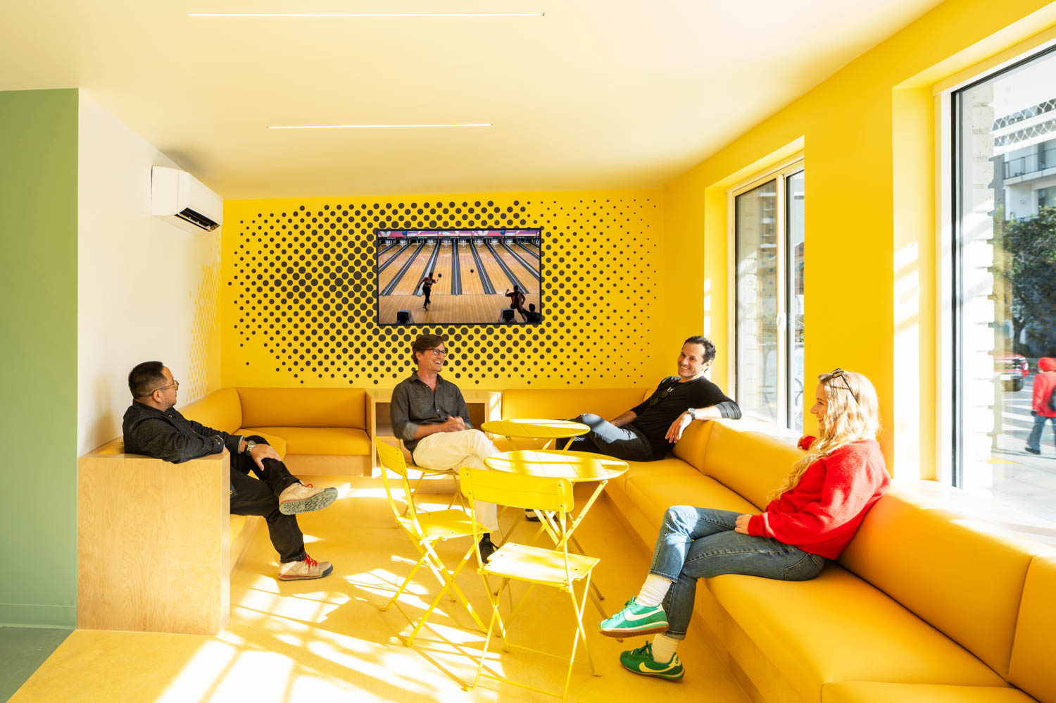 Four people sit on yellow couches and chairs in a bright, yellow room with large windows. A television on the wall displays a bowling alley scene.