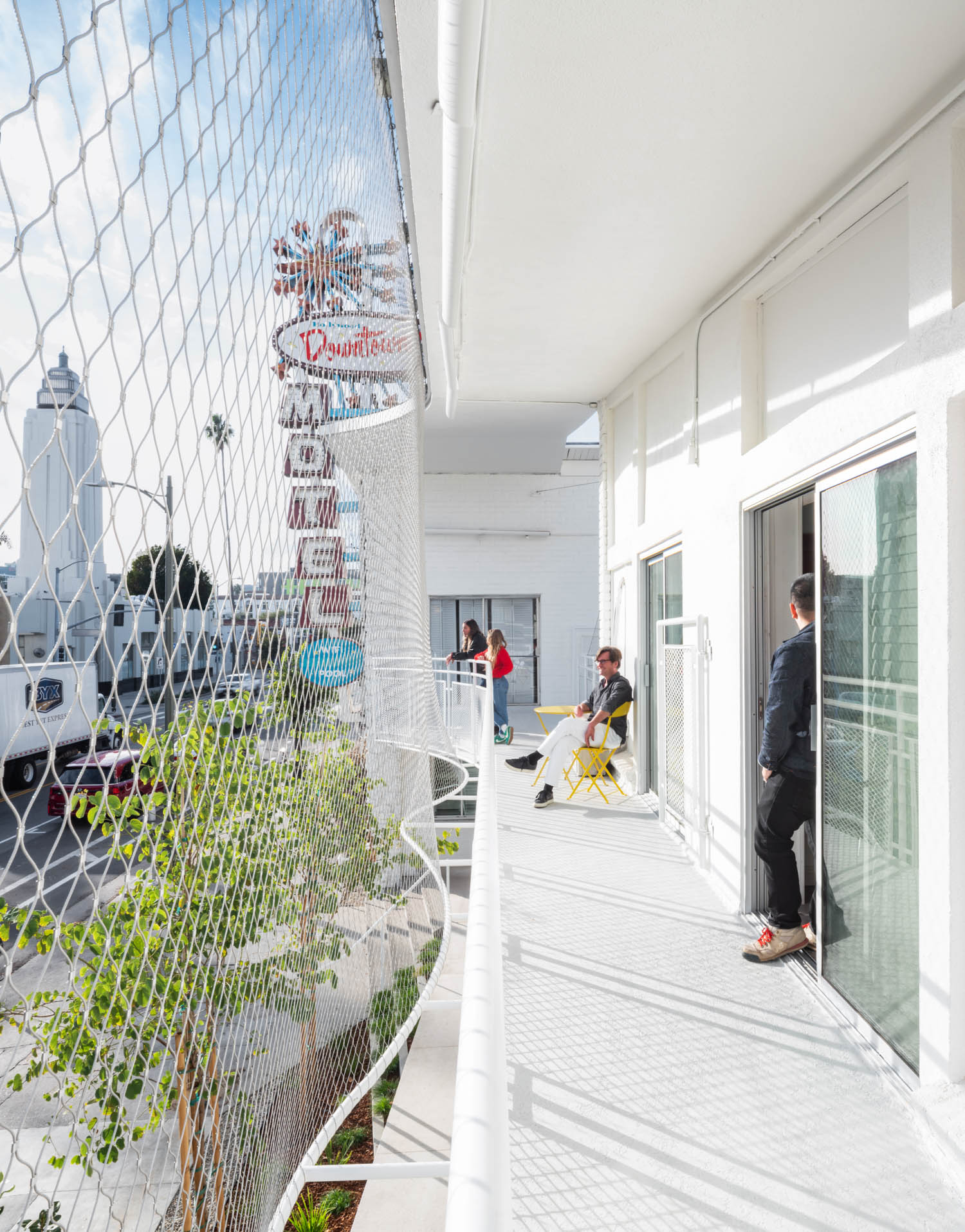 People stand and sit on a white balcony outside a modern building, with a city street and a sign reading "Downtown" visible in the background. A netting covers the balcony rail.