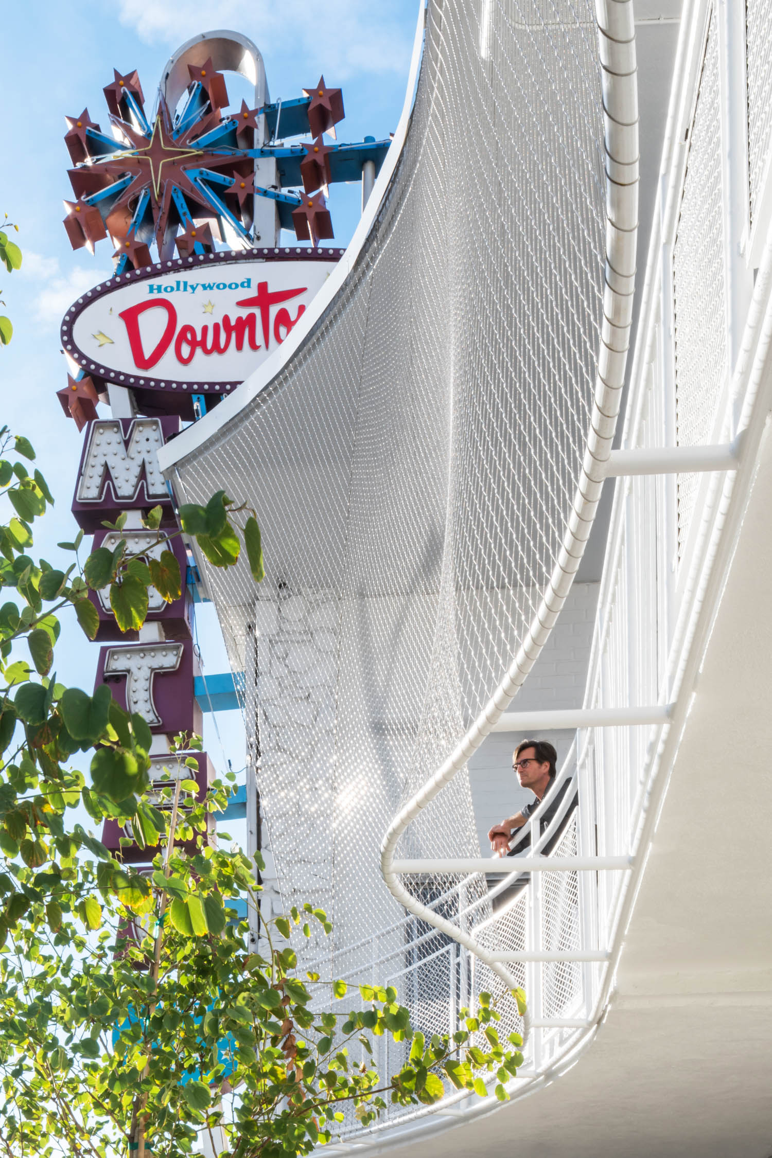 A man stands on a white motel balcony near a retro sign reading "Hollywood Downtowner Motel," with trees partially obscuring the view.