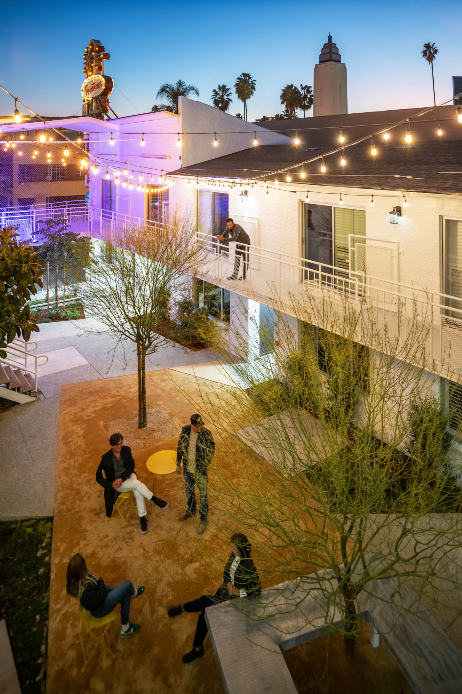 People sit and stand in a courtyard of a modern building at dusk, with string lights overhead and palm trees in the background.
