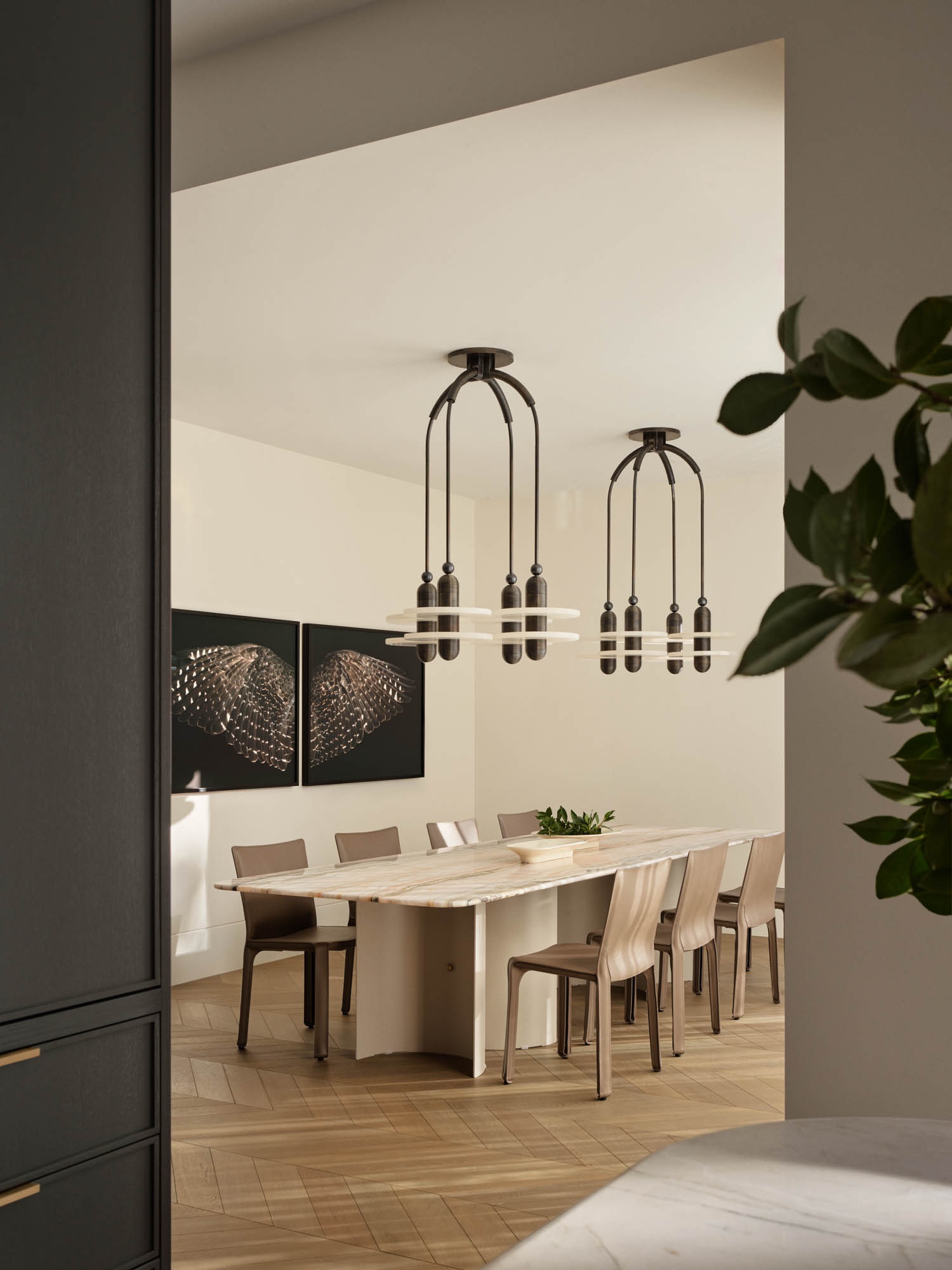 Modern dining room with a long table, wooden chairs, two pendant light fixtures, minimalist decor, and abstract art on the wall. A leafy plant is visible in the foreground.