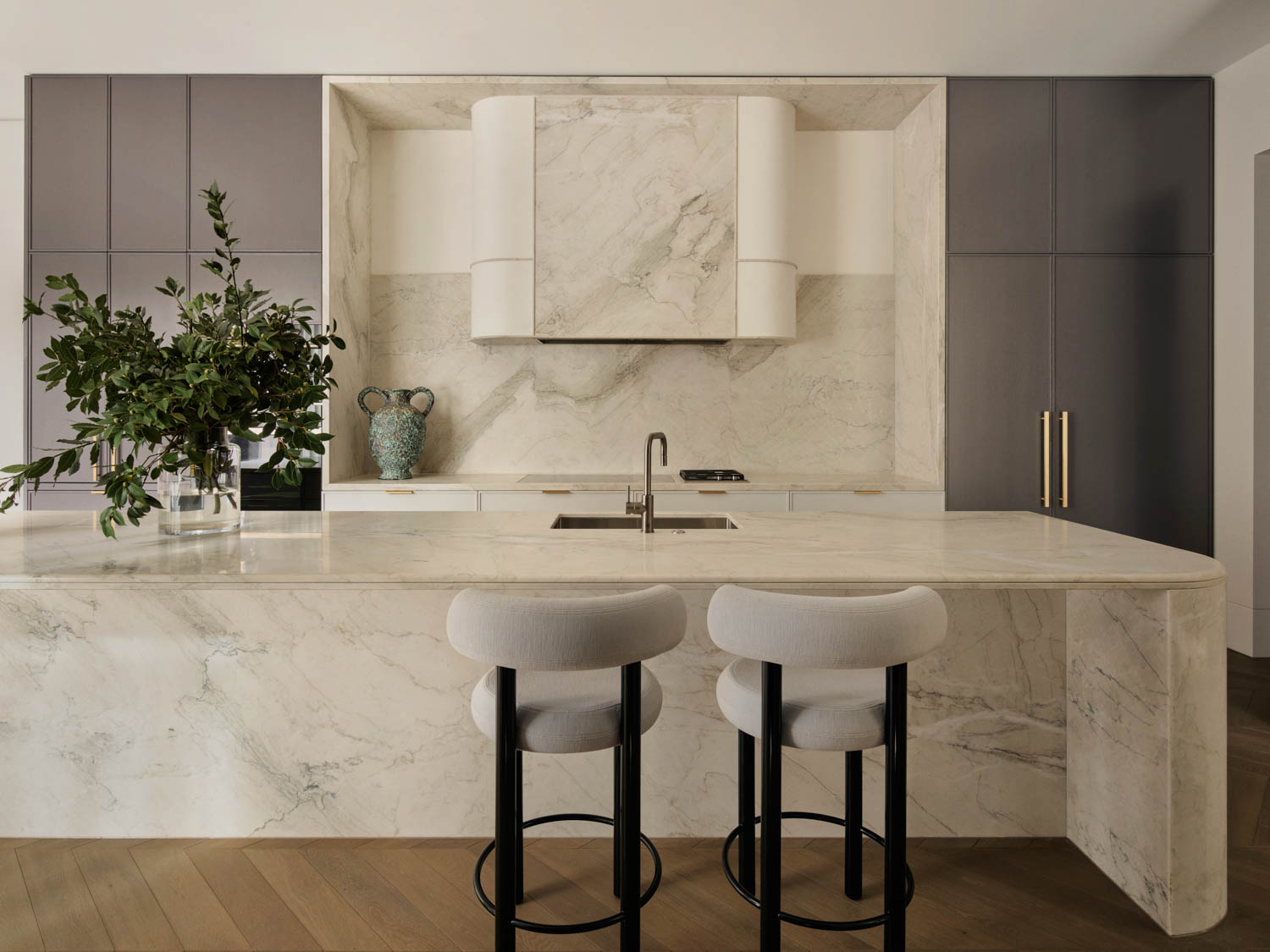 Modern kitchen with marble countertops and backsplash, two cushioned bar stools, a sink, built-in stovetop, dark cabinets, and decorative plants on the island.